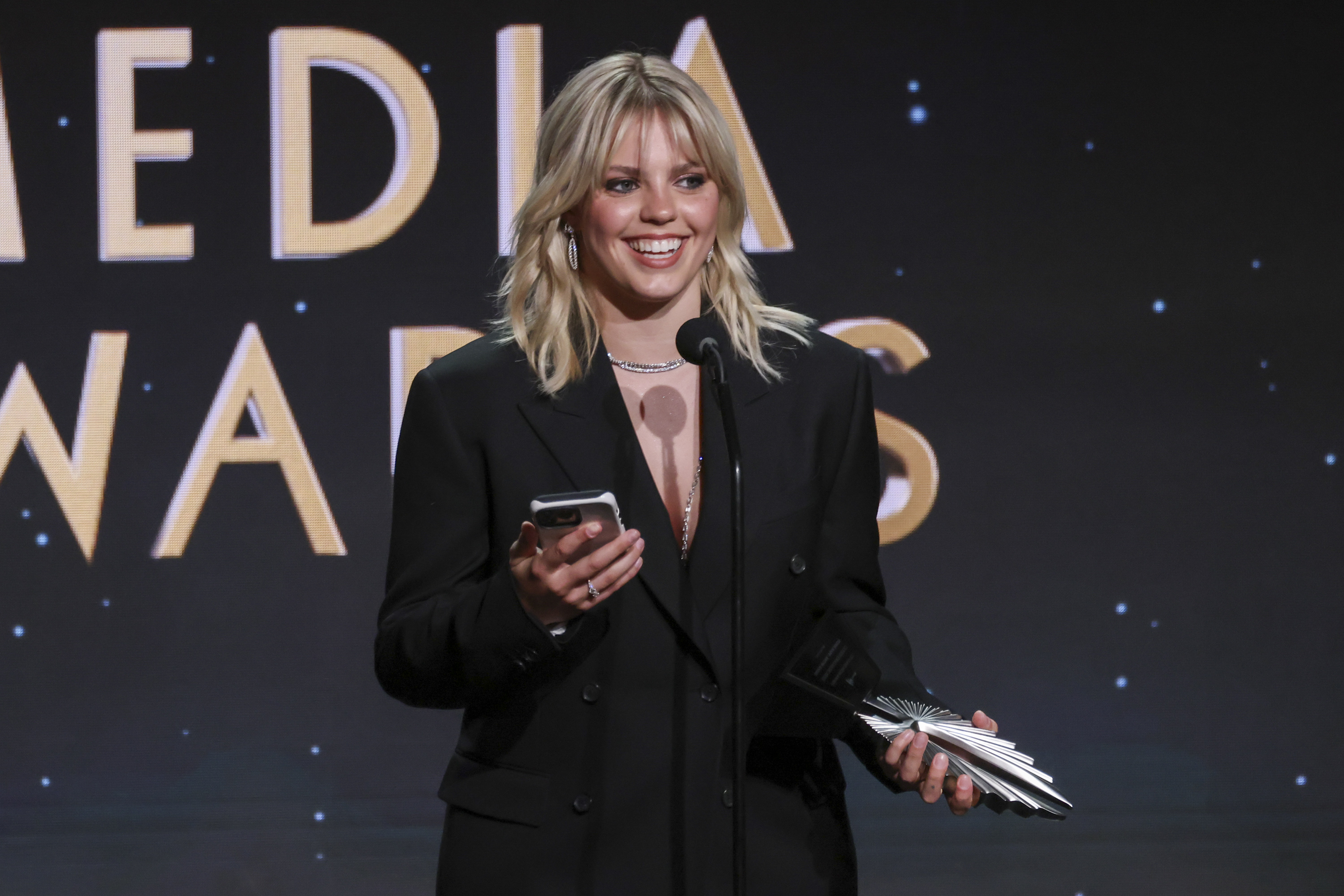 Reneé in a blazer holding a phone and award at a podium with &quot;MEDIA AWARDS&quot; in the background