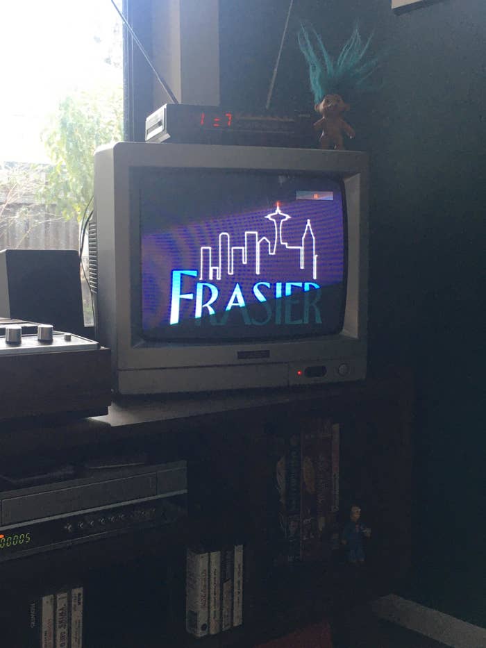 Old CRT TV displaying &quot;Frasier&quot; title screen, on a cabinet with gaming console, dolls nearby