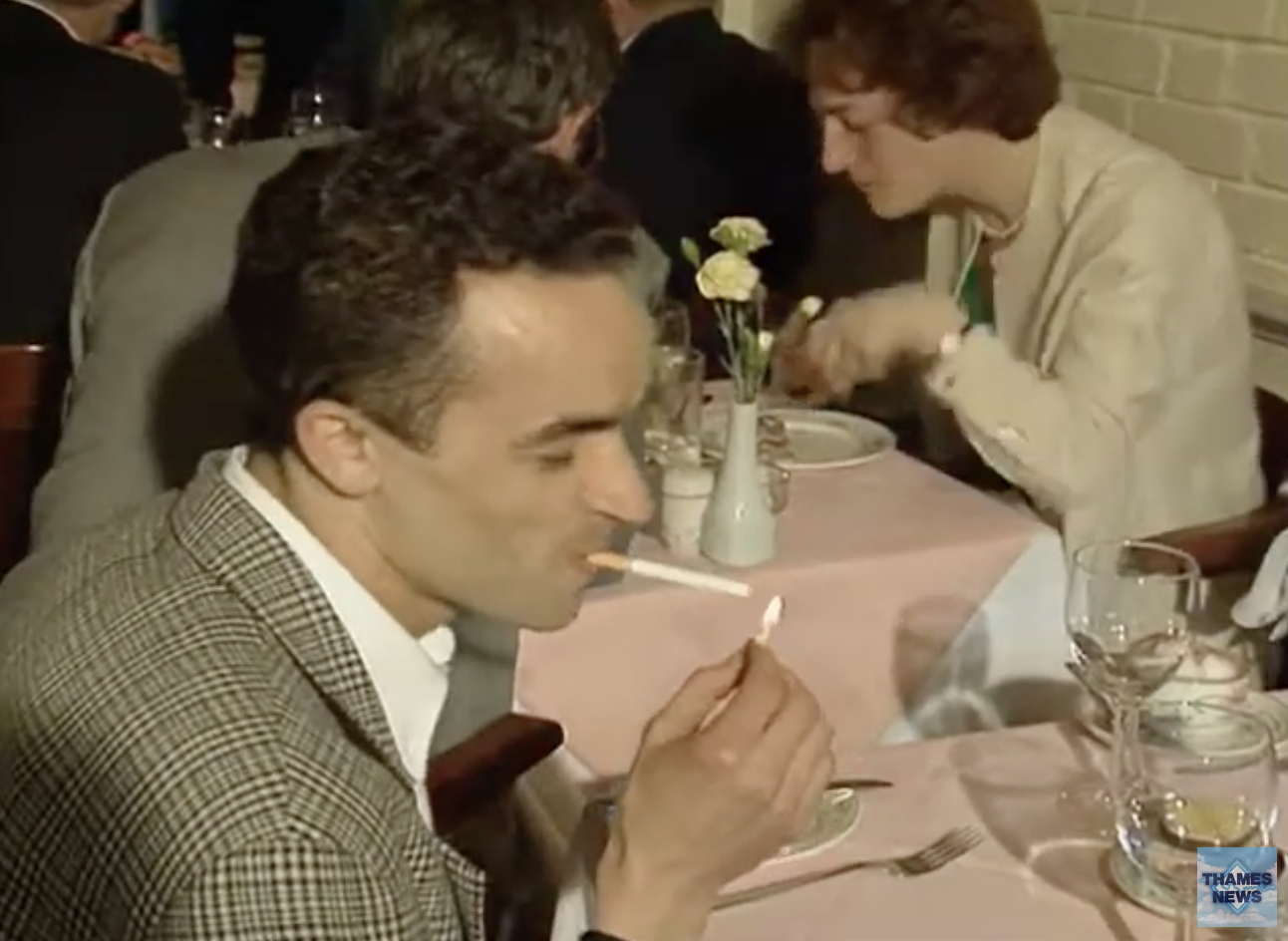 Man in a checkered jacket lighting a cigarette at a restaurant, with another patron in the background