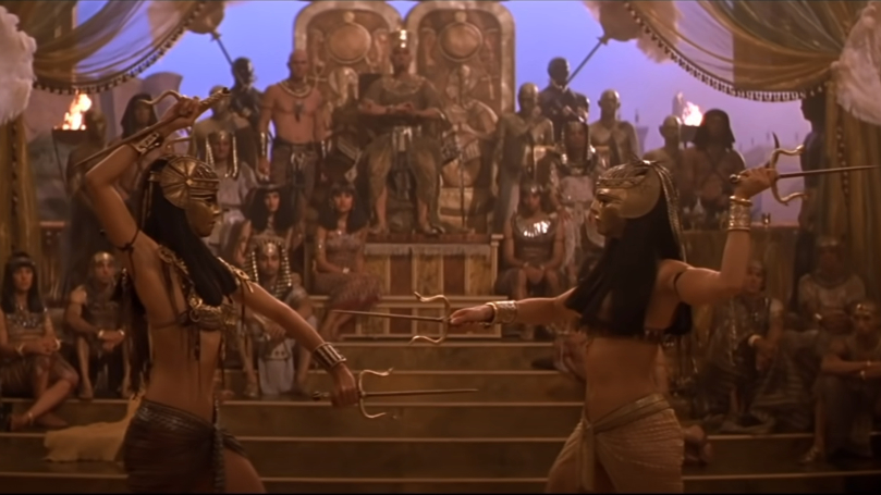 Two characters in ancient Egyptian style attire perform about to strike in a grand hall filled with onlookers