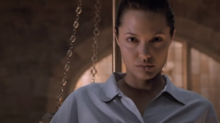 Woman with hair pulled back, wearing a buttoned shirt, standing in a dimly lit room with chains