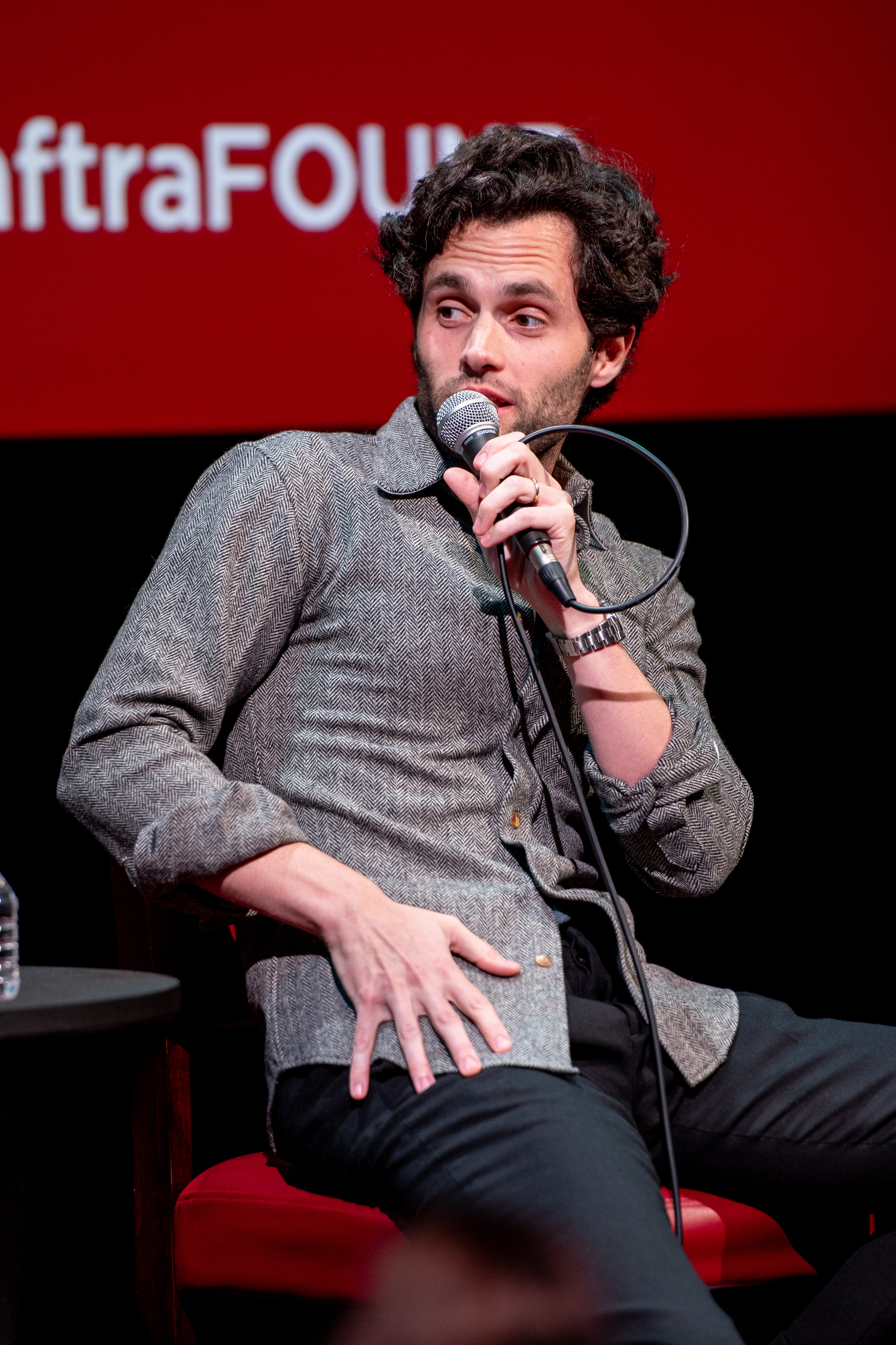 Penn Badgley seated with a microphone, wearing a buttoned shirt and jacket, at a speaking event
