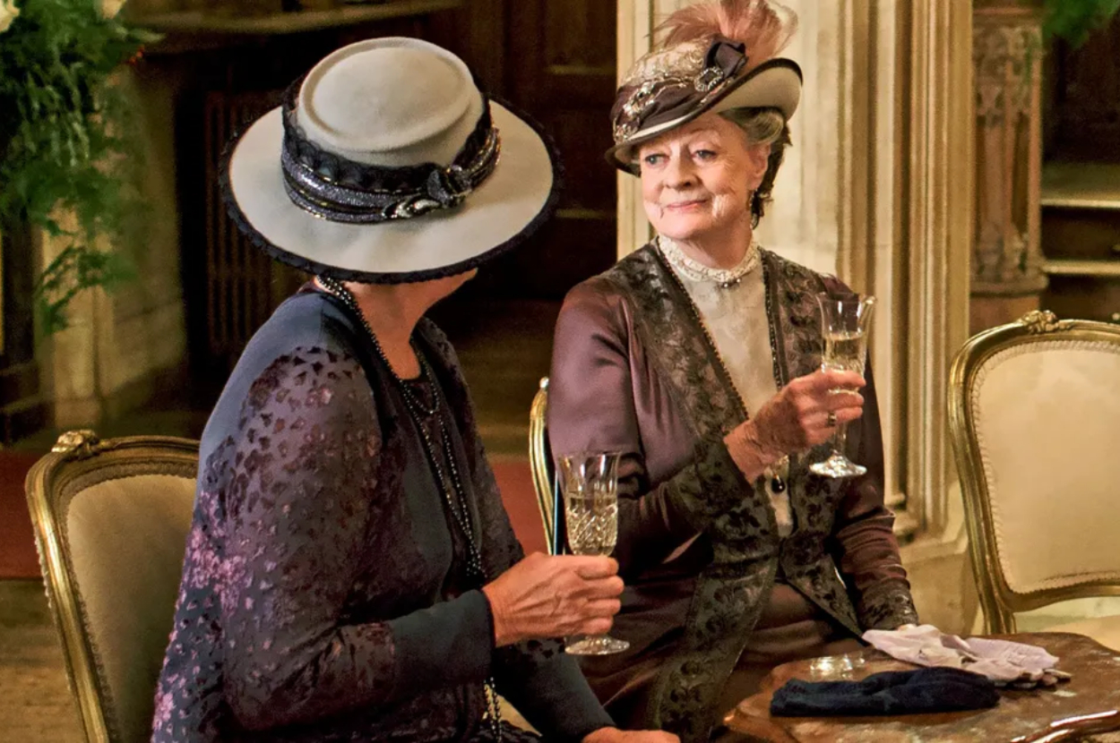 Two characters from Downton Abbey in early 20th-century attire, seated with drinks. Victorian decor surrounds them
