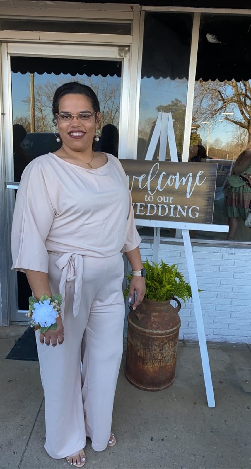 Woman in elegant beige jumpsuit with corsage poses next to &#x27;Welcome to our wedding&#x27; sign