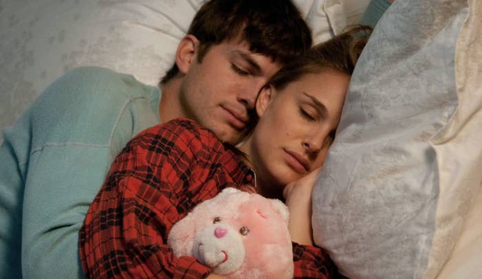 Two actors portraying a couple asleep in bed, cuddling a pink teddy bear