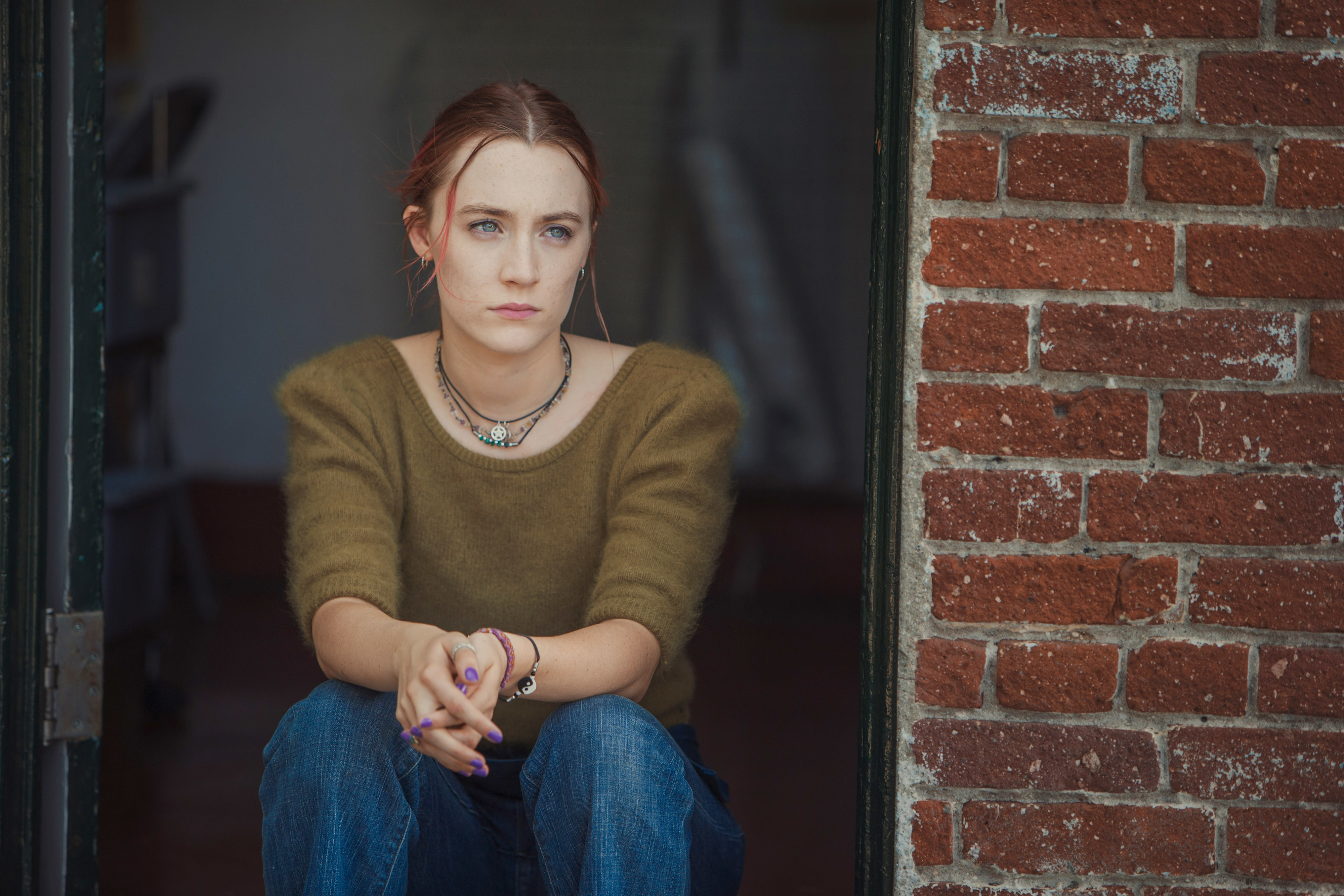 Lady Bird sitting by a window, looking pensive, wearing a sweater and a necklace