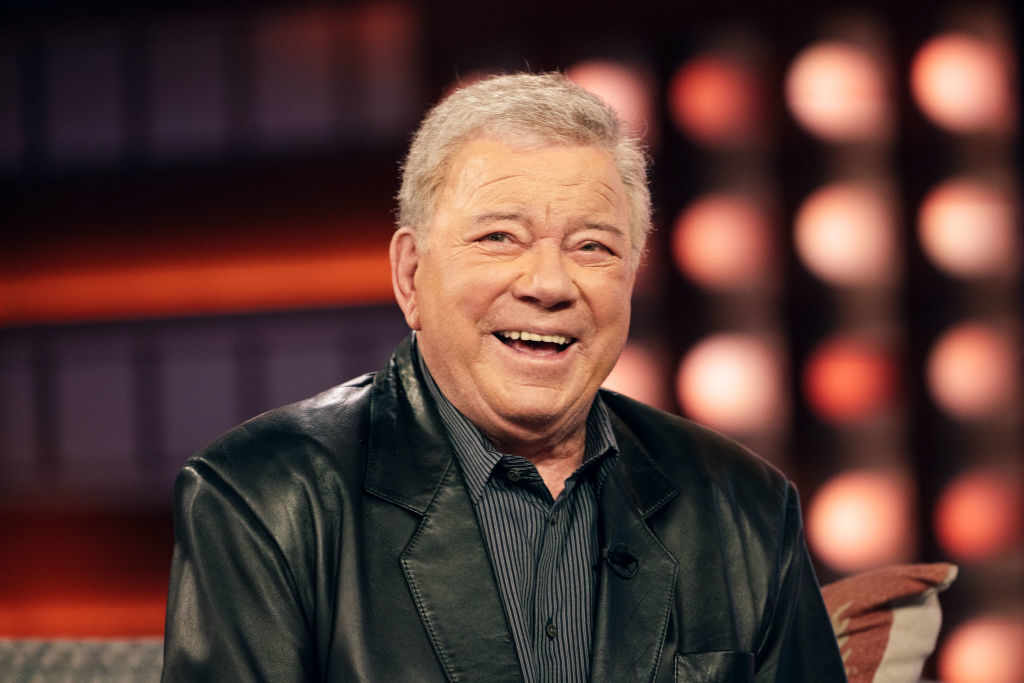 Smiling person wearing a jacket, sitting in a talk show environment with blurred lights in the background