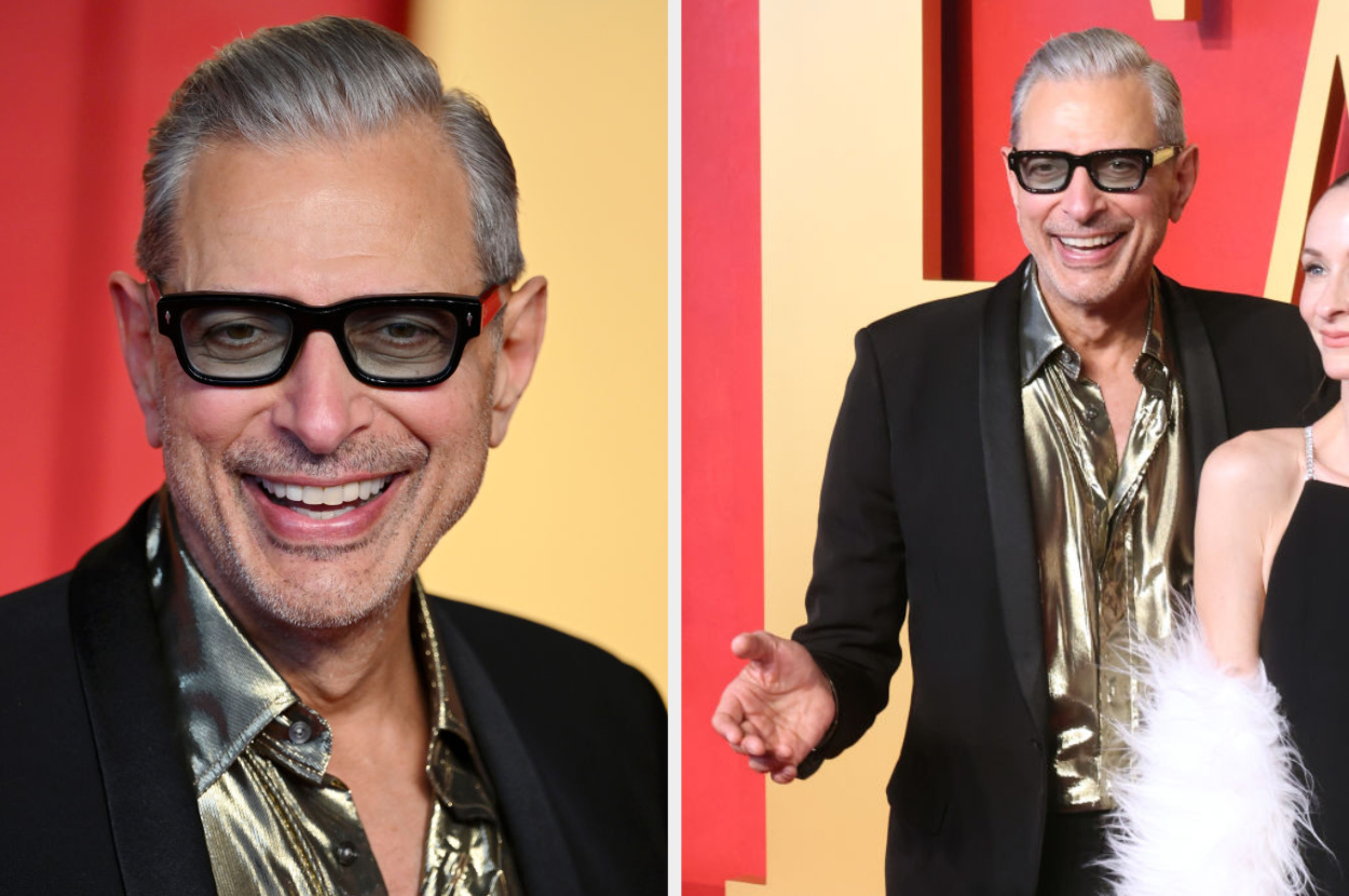 Jeff Goldblum on the red carpet, wearing glasses and a black suit with a metallic shirt, alongside a person in a feathered outfit