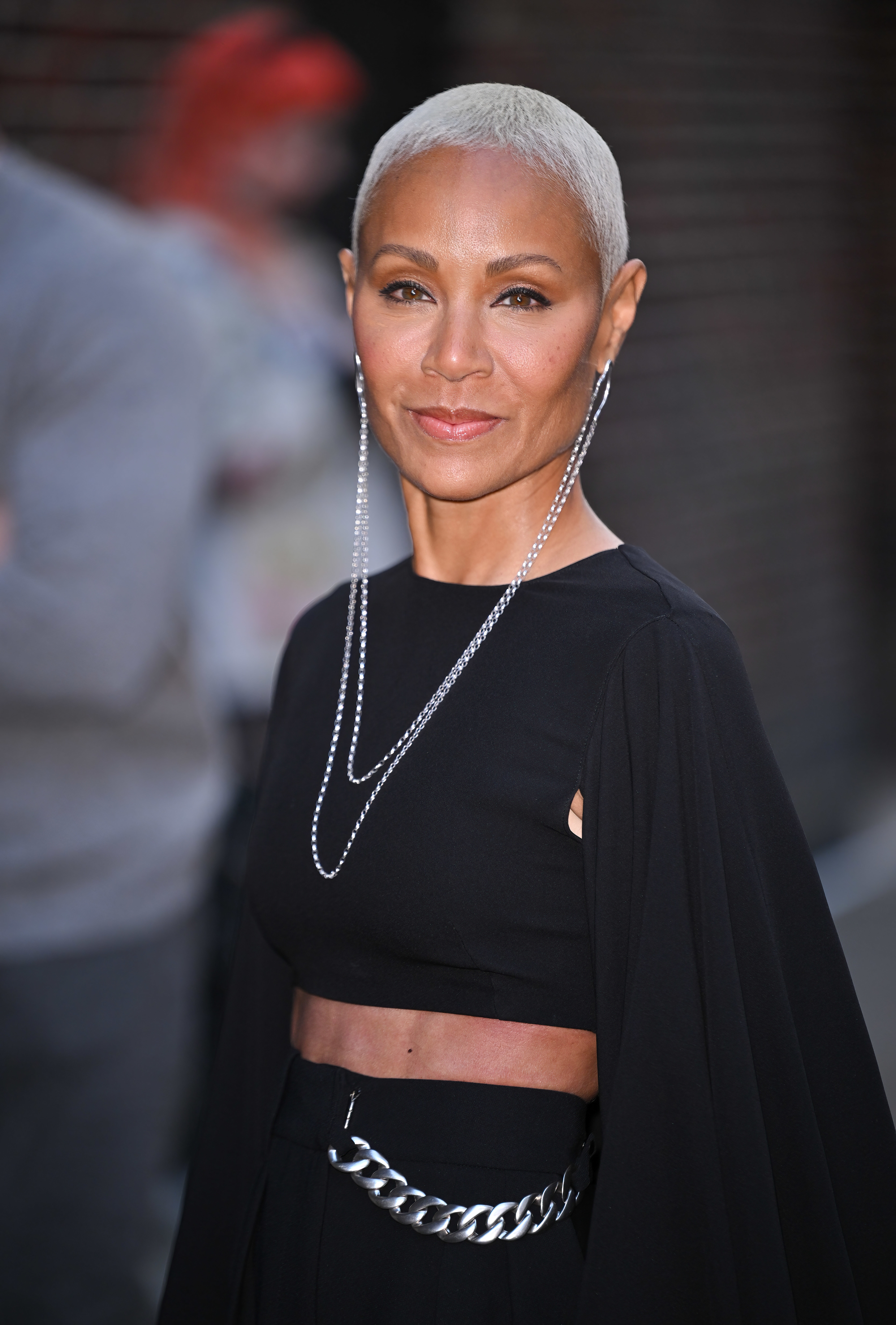 Jada wearing a black outfit with a chain belt and draped sleeves, at an event