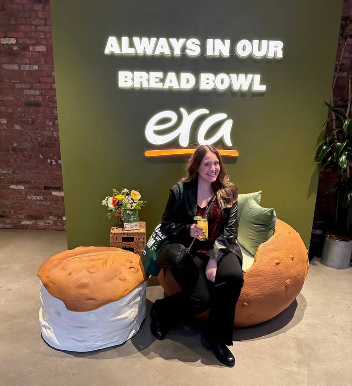 Woman sitting on a bread bowl-shaped chair at an &quot;ALWAYS IN OUR BREAD BOWL ERA&quot; event