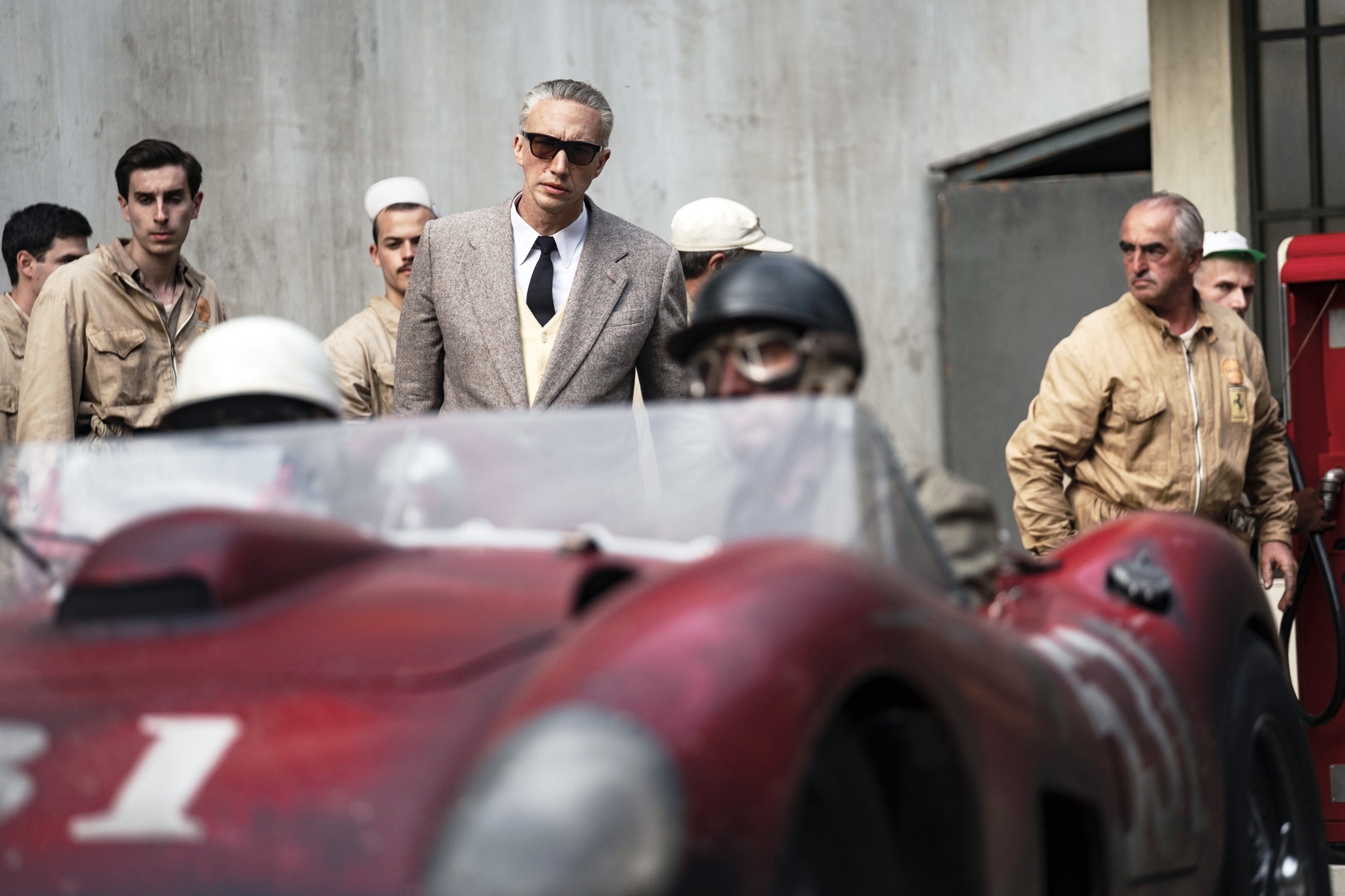 Enzo Ferrari in 1950s attire stands confidently among vintage race cars and pit crew
