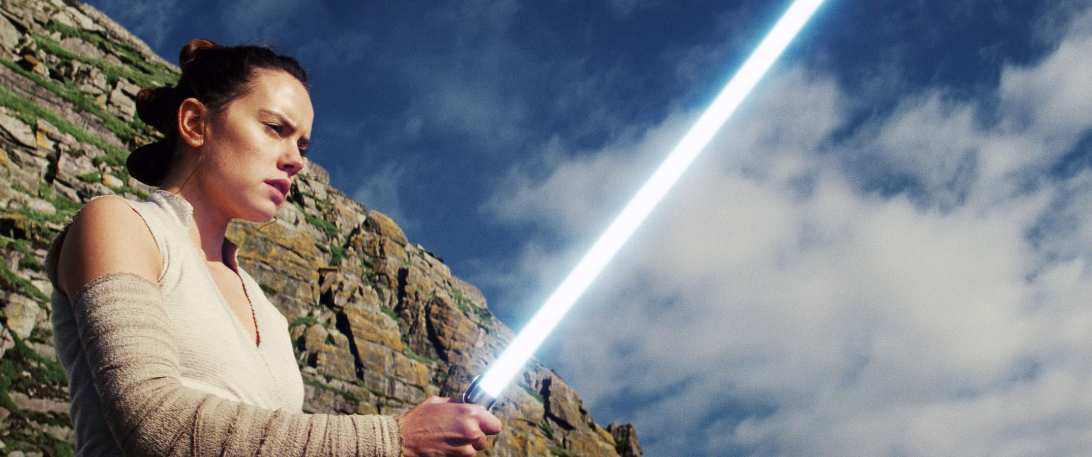 Rey from Star Wars holds a lightsaber on a rocky terrain, looking focused