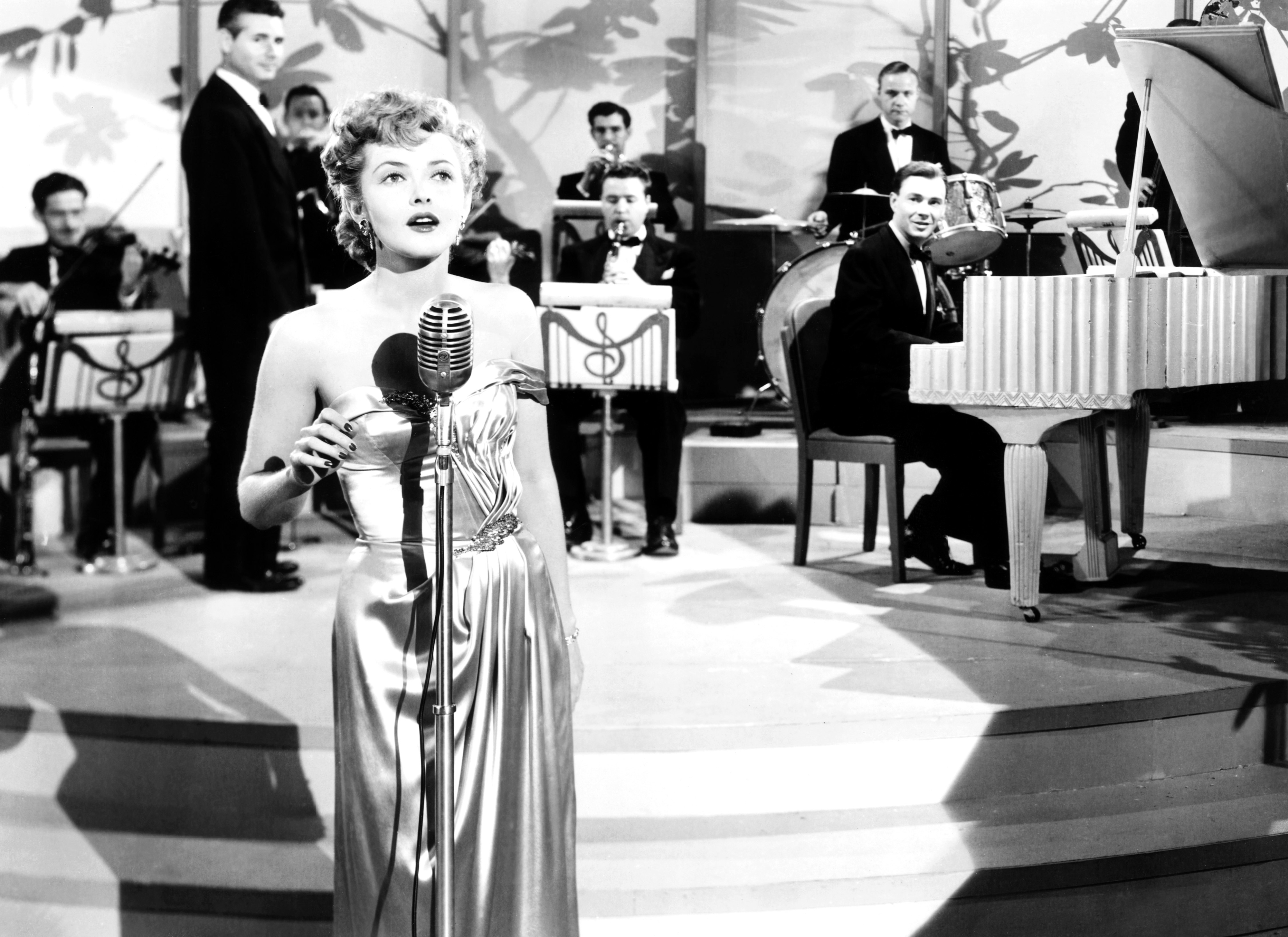 Kitty in elegant dress sings into vintage microphone on stage, band with pianist in background