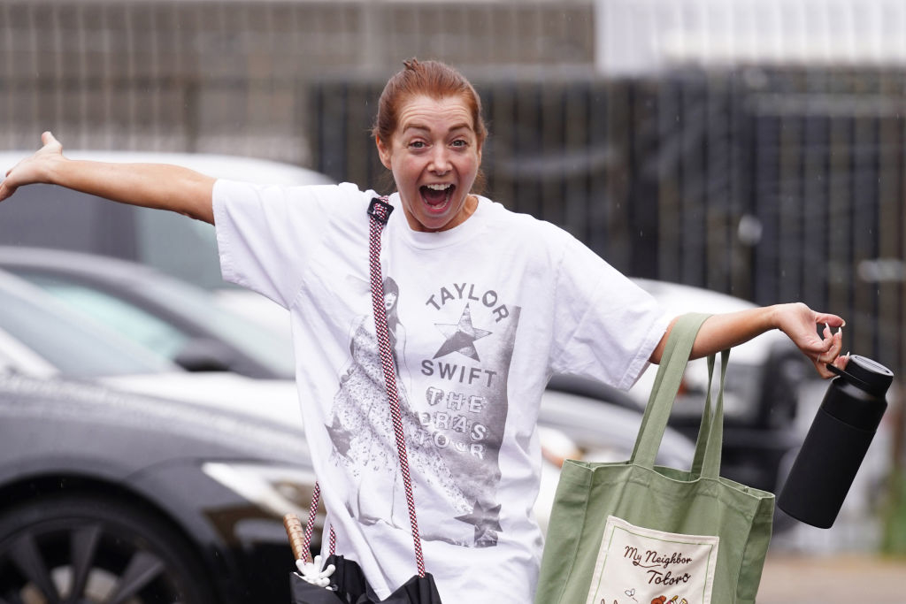 Woman in a Taylor Swift T-shirt with open arms and an excited expression, carrying a tote bag