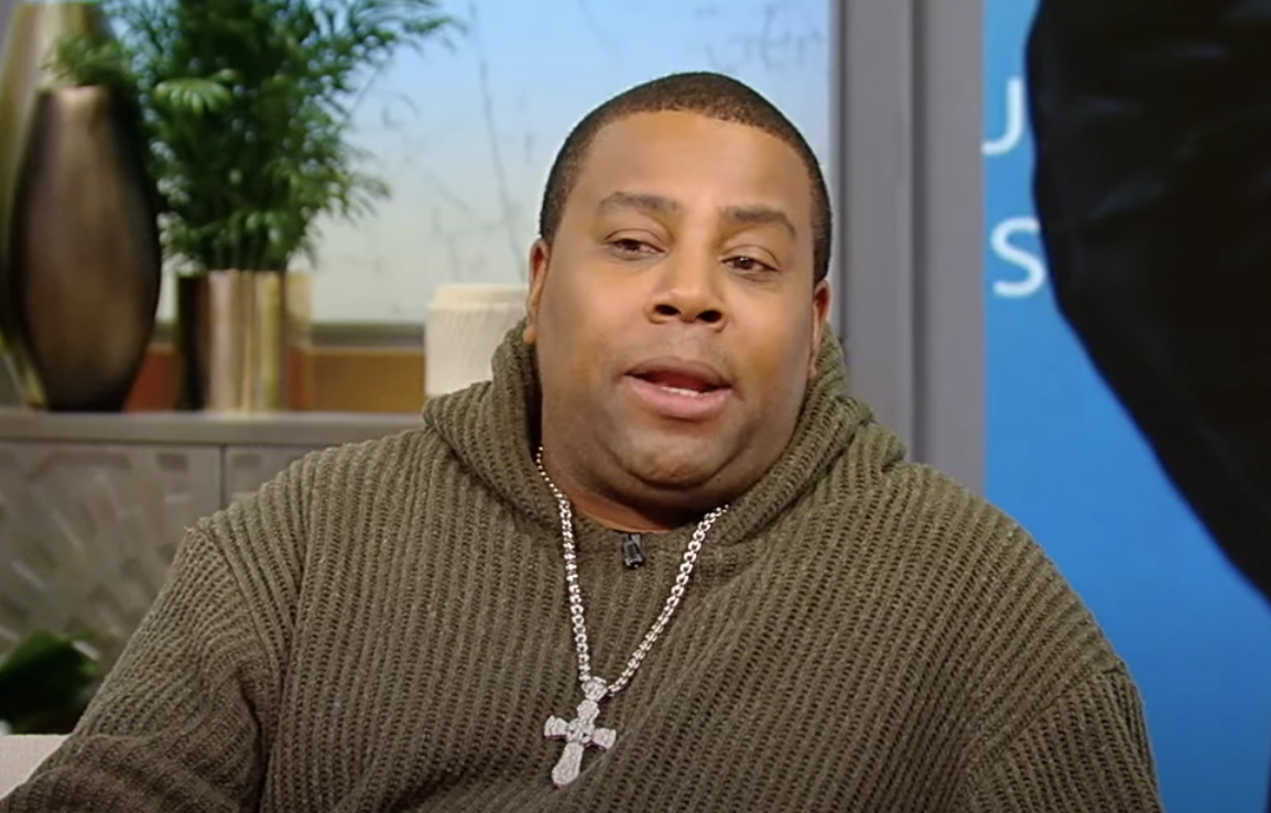 Kenan in sweater, wearing chain with cross pendant, appears to speak in an interview setting