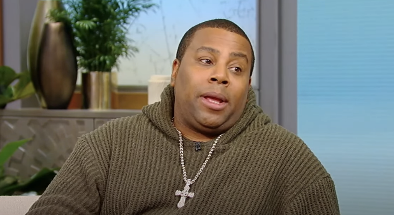 Kenan in a knit sweater with a cross necklace, sitting indoors, mid-conversation