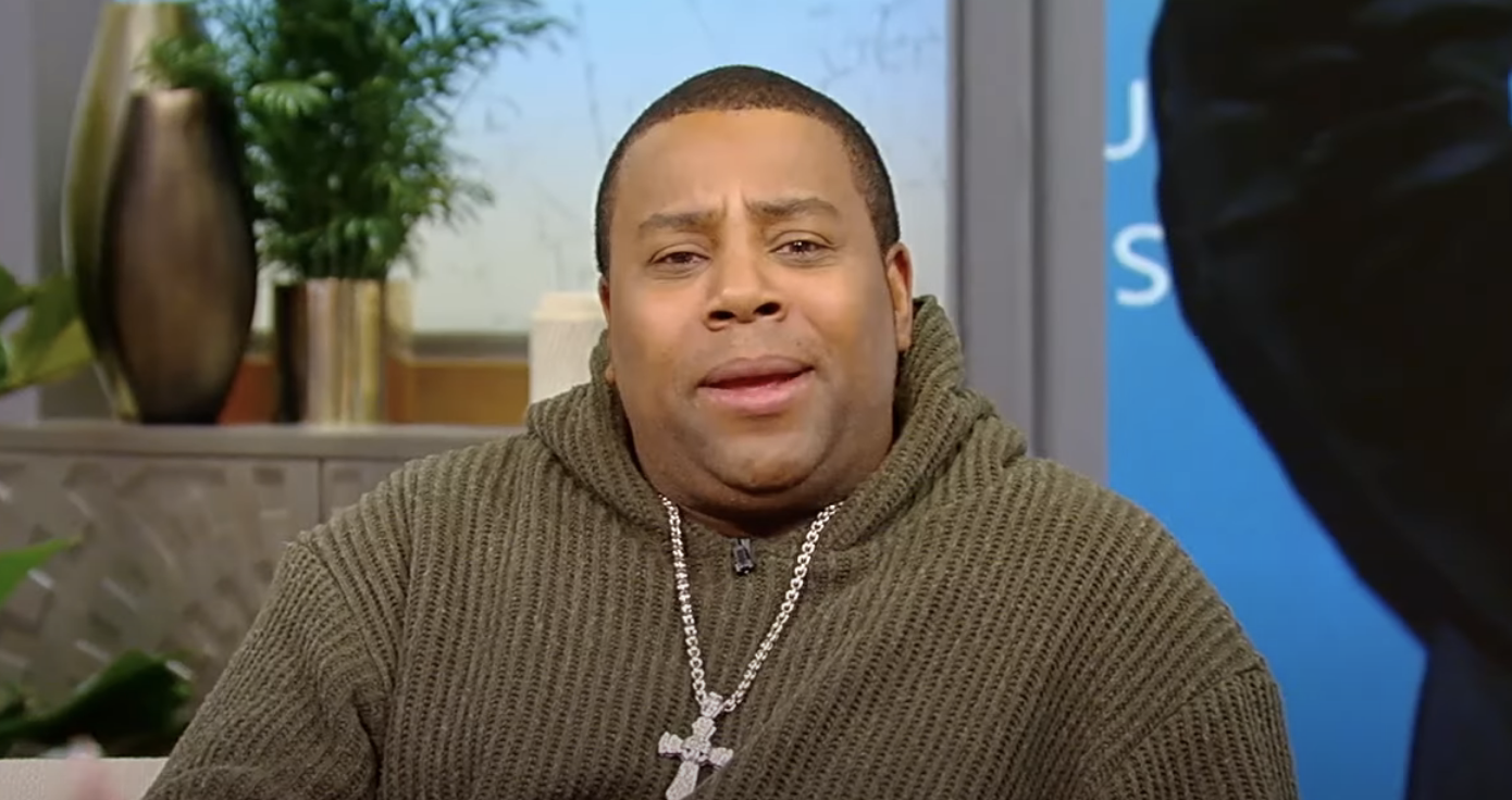 Kenan  in sweater and necklace with an angry expression on a talk show set