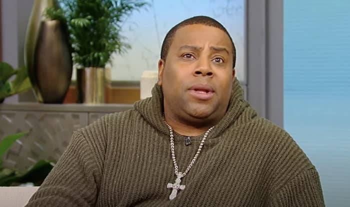 Kenan with a surprised expression wearing a knit sweater and a cross necklace