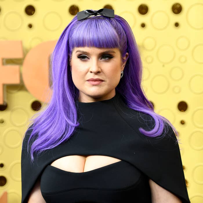 Kelly with purple hair and bow, wearing a dark outfit with a cutout detail