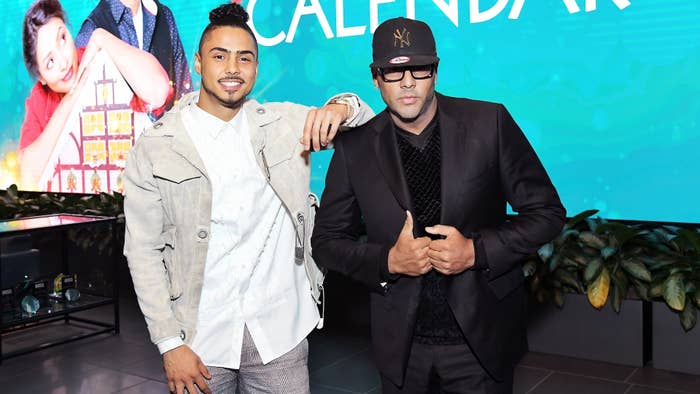 Two men posing for a photo with promotional signage in the background. The man on the left is wearing a casual suit, and the one on the right is in a black outfit with a cap