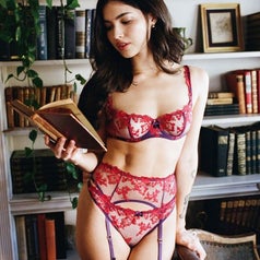 Woman in lace lingerie set with floral patterns, posing with an open book in a library setting