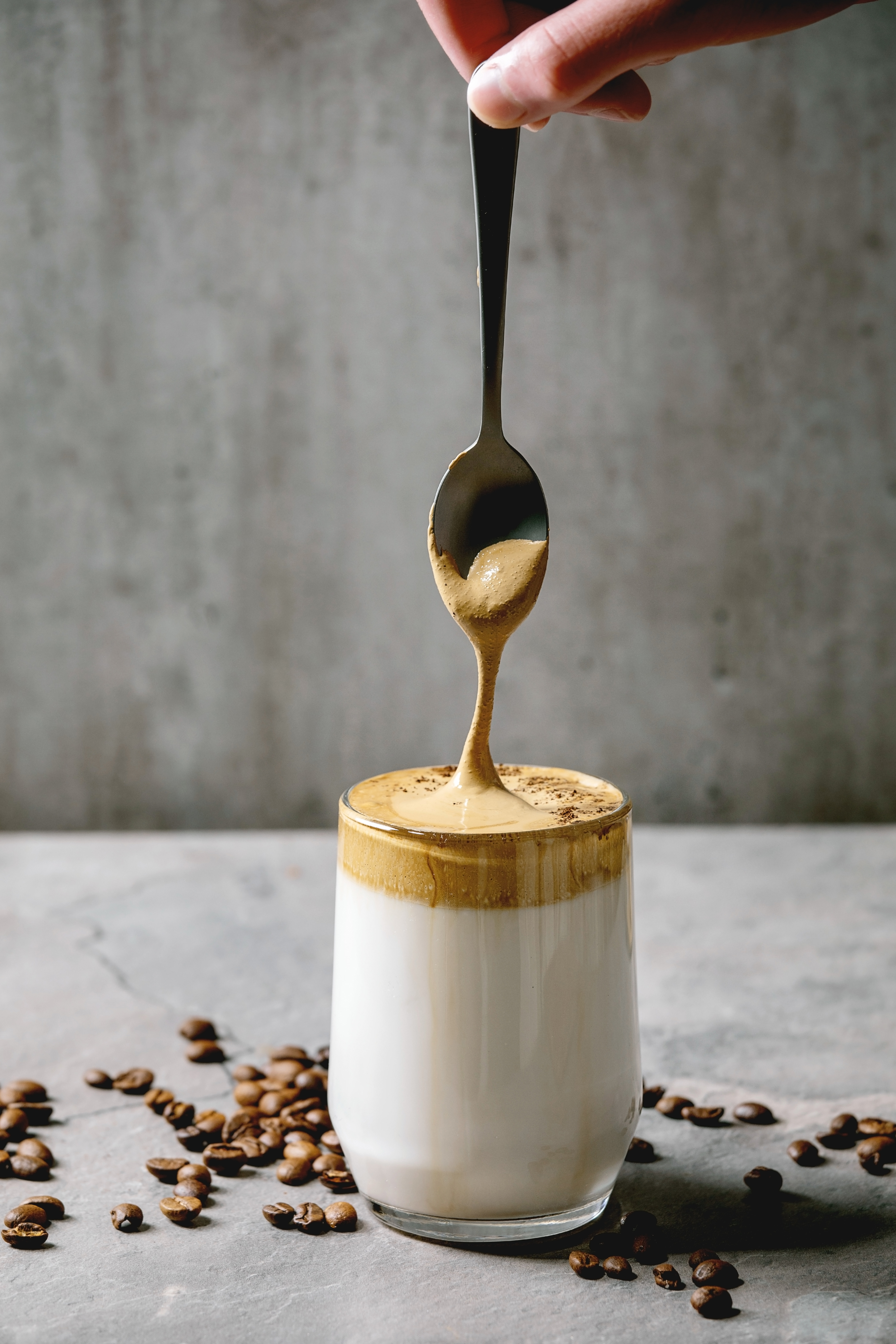 Person&#x27;s hand drizzling whipped coffee over milk in a glass, surrounded by scattered coffee beans