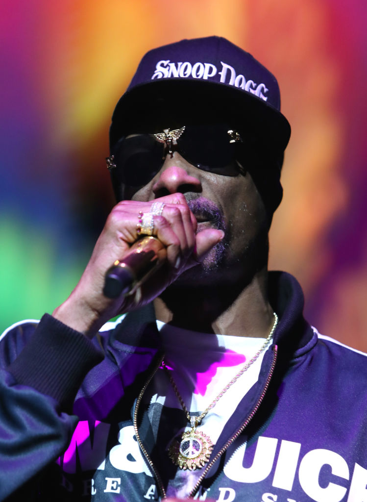 Snoop Dogg performing, wearing sunglasses, a hat, necklace, and rings, holding a microphone