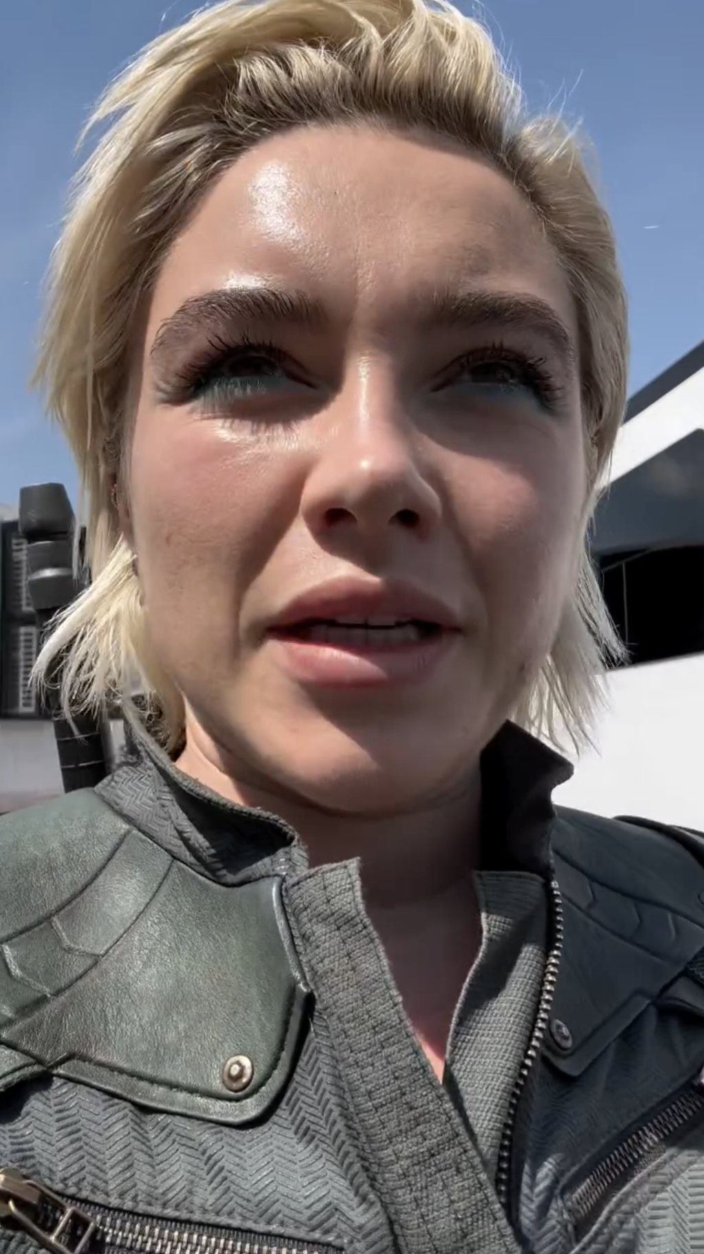 Florence Pugh with short blonde hair in a leather jacket, looking into the camera, with a bright sky behind