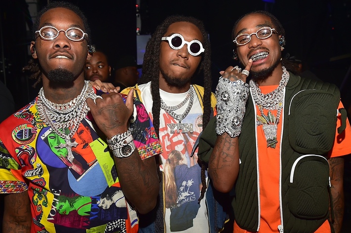 Three men posing together, two wearing glasses and abundant jewelry. The one in the middle has a graphic tee and vest