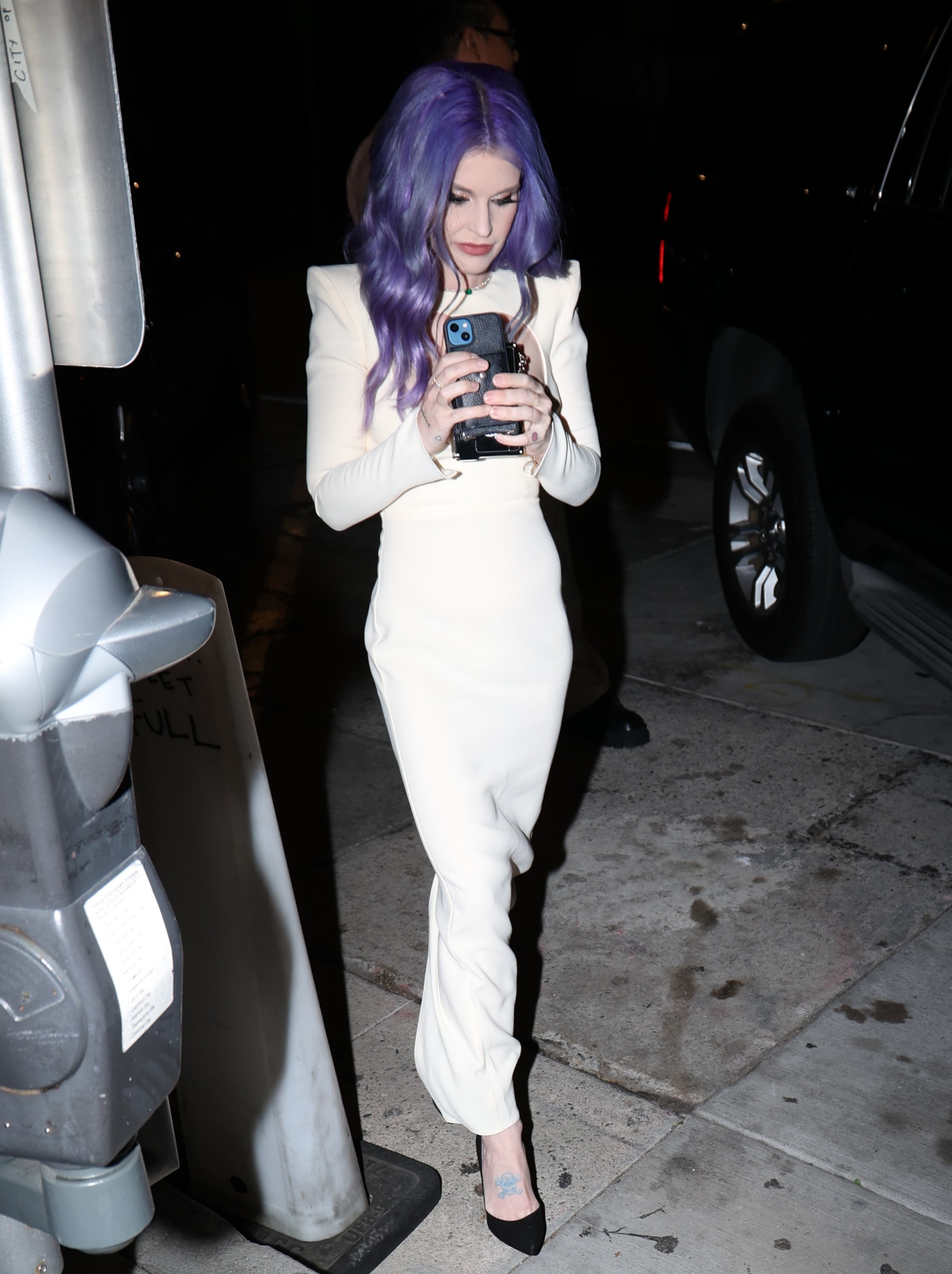Kelly with purple hair in a white outfit using her phone on a city street at night