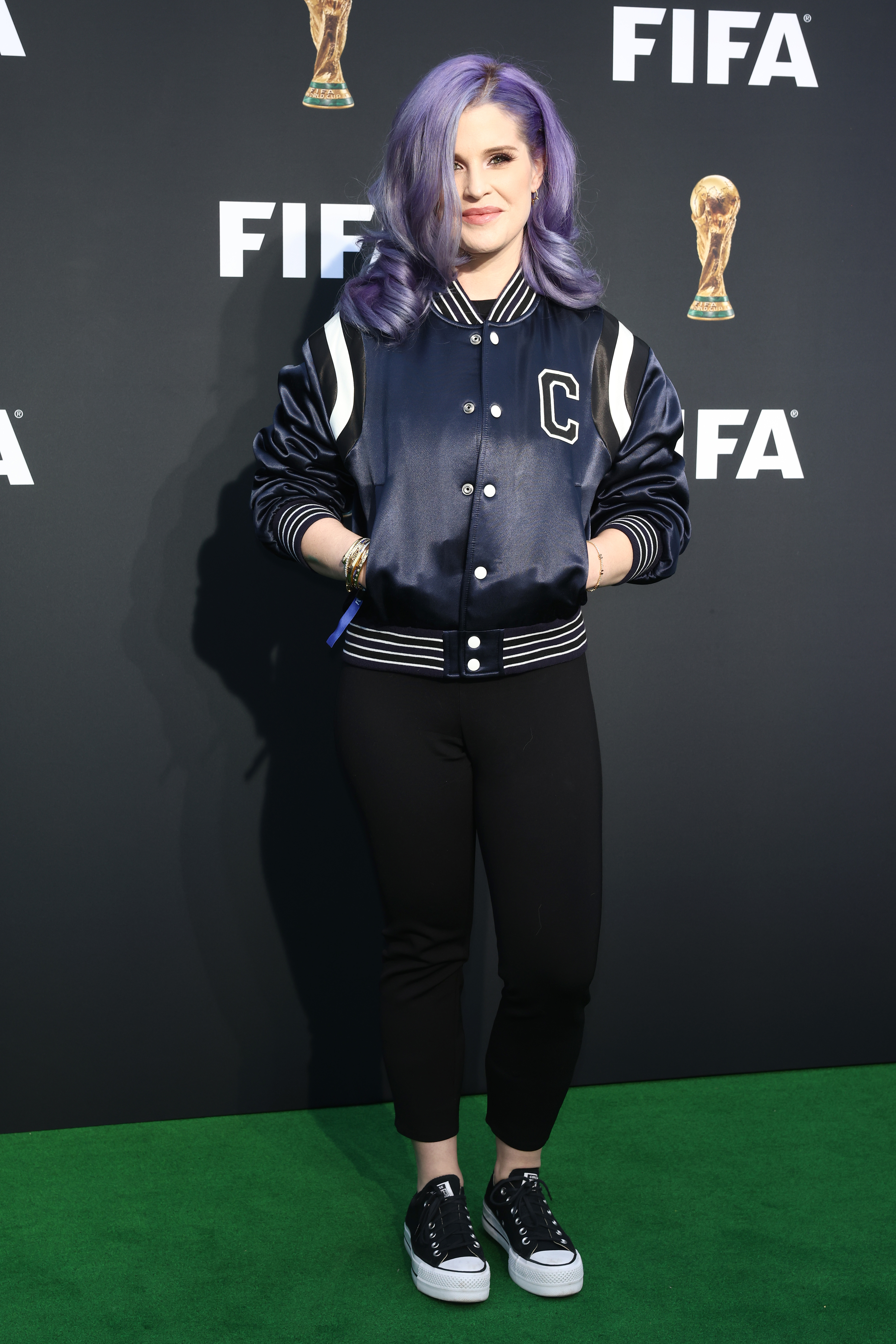 Kelly in a varsity jacket and pants standing on an event carpet