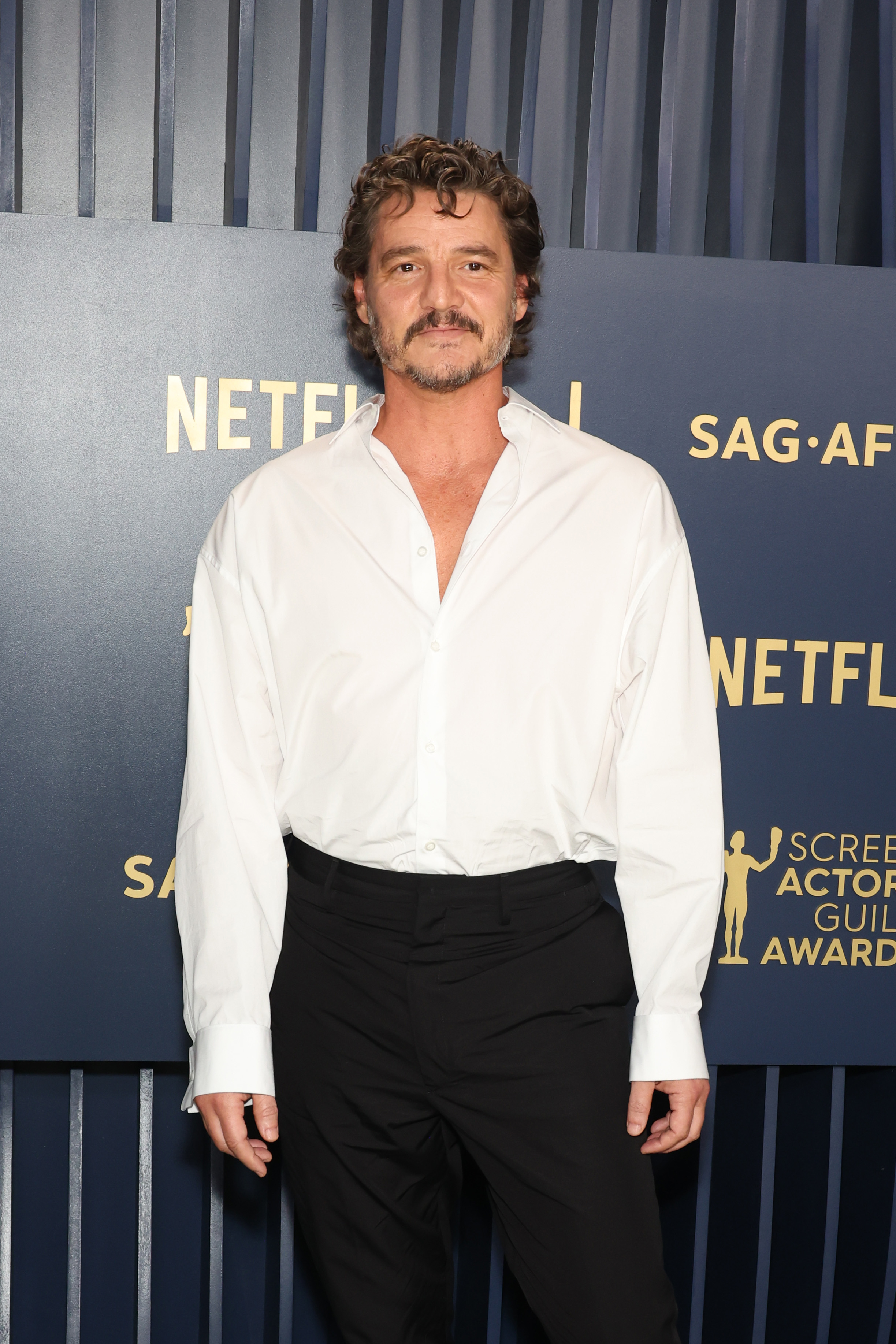 Pedro Pascal stands smiling in a semi-formal outfit with an unbuttoned white shirt and black trousers at a press event