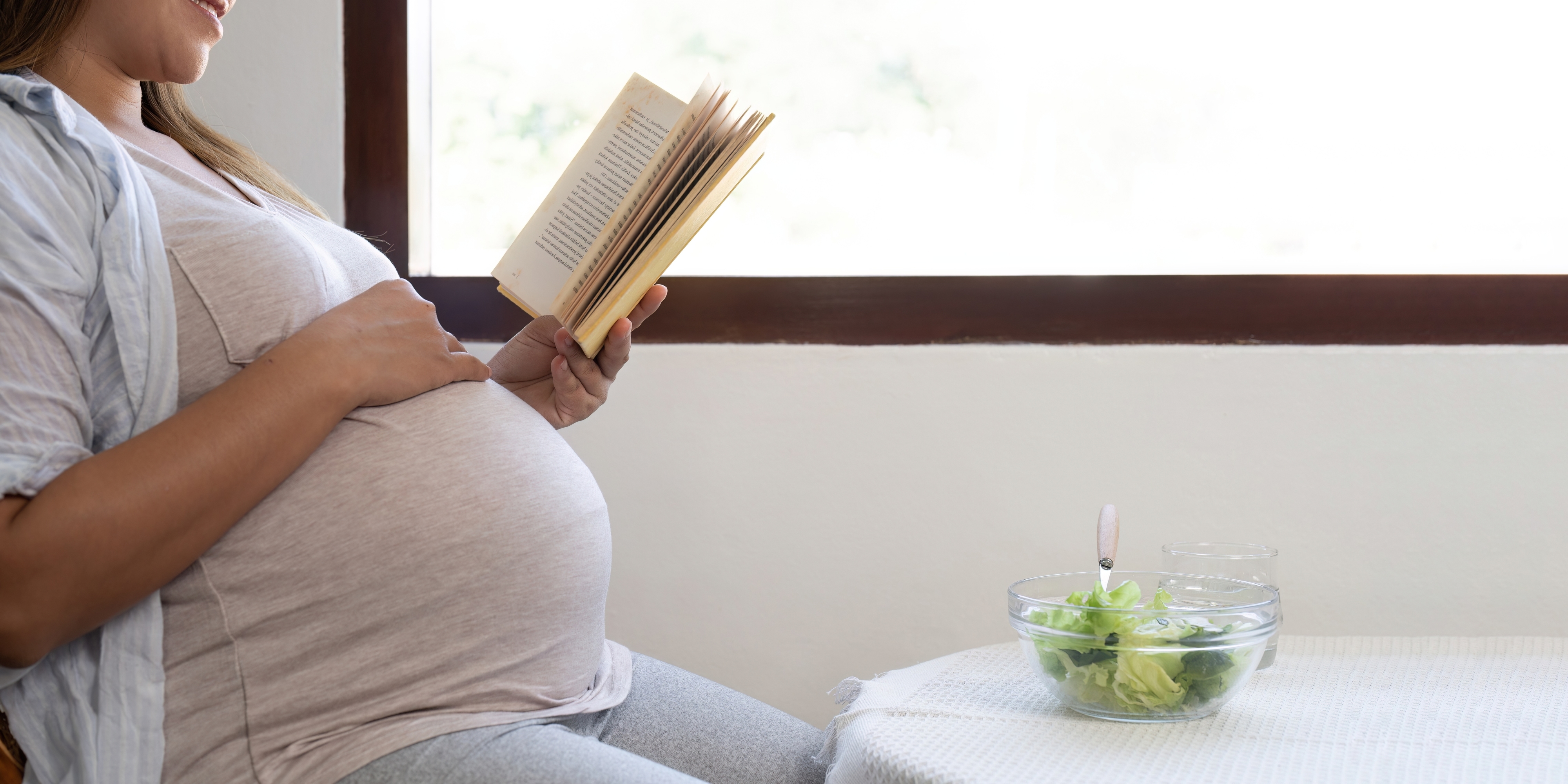 Pregnant person sitting, reading a book, with a salad on the table next to them