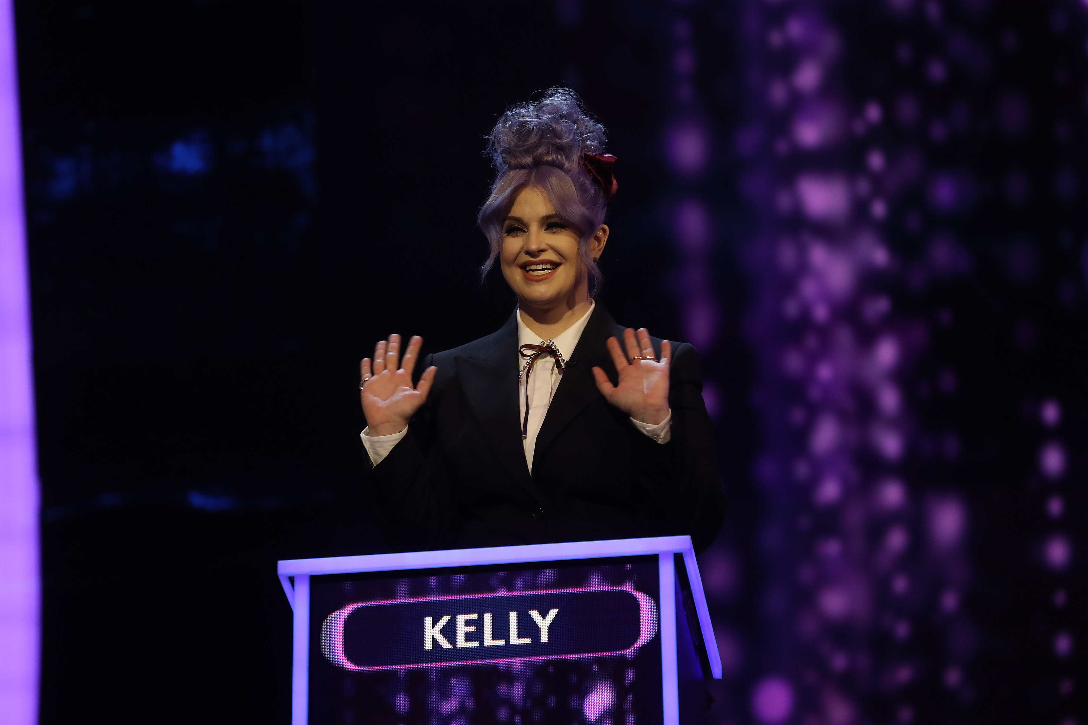 Kelly smiling and waving on a game show set standing behind a podium, dressed in a suit with a unique hairstyle