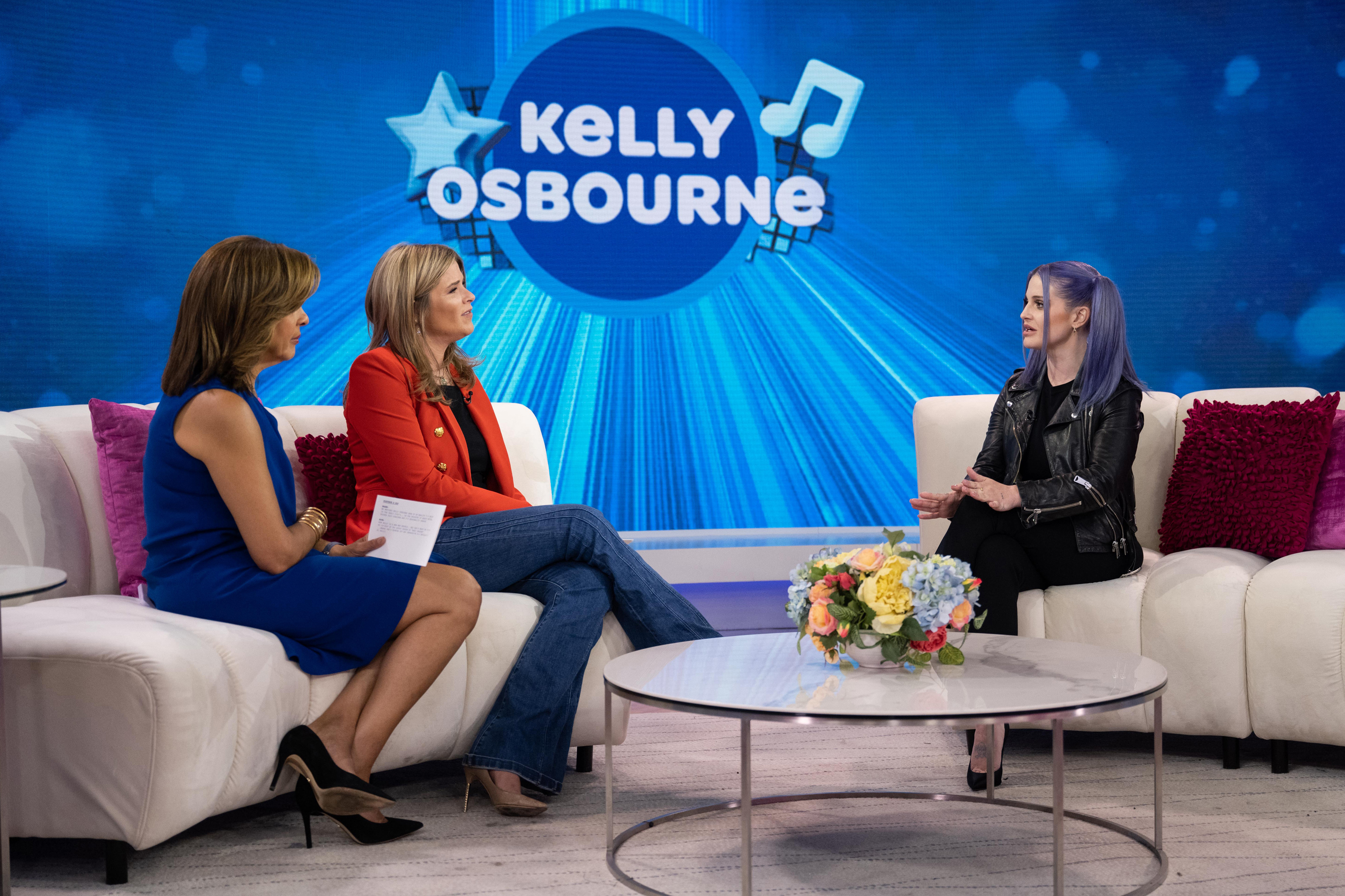 Kelly Osbourne chats with Hoda Kotb and Jenna Bush Hager on Today show, seated on a couch, all in front of a show-themed backdrop