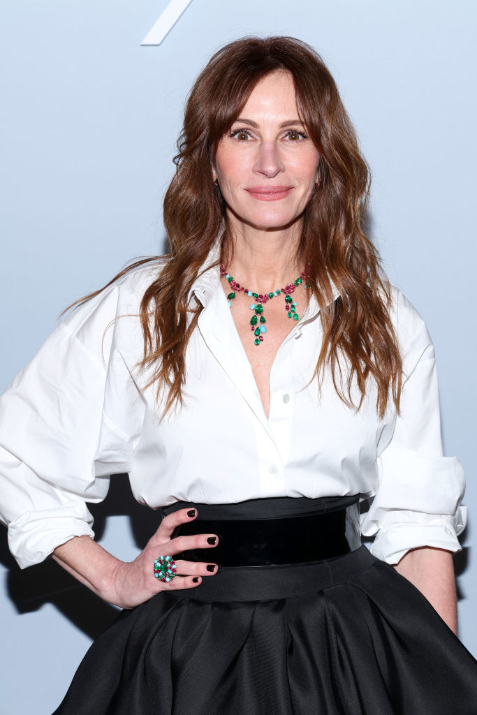 Julia Roberts posing, wearing a white blouse, black belt, and black skirt, accessorized with a green necklace and ring