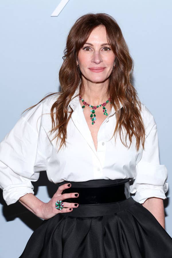 Julia Roberts posing, wearing a white blouse, black belt, and black skirt, accessorized with a green necklace and ring