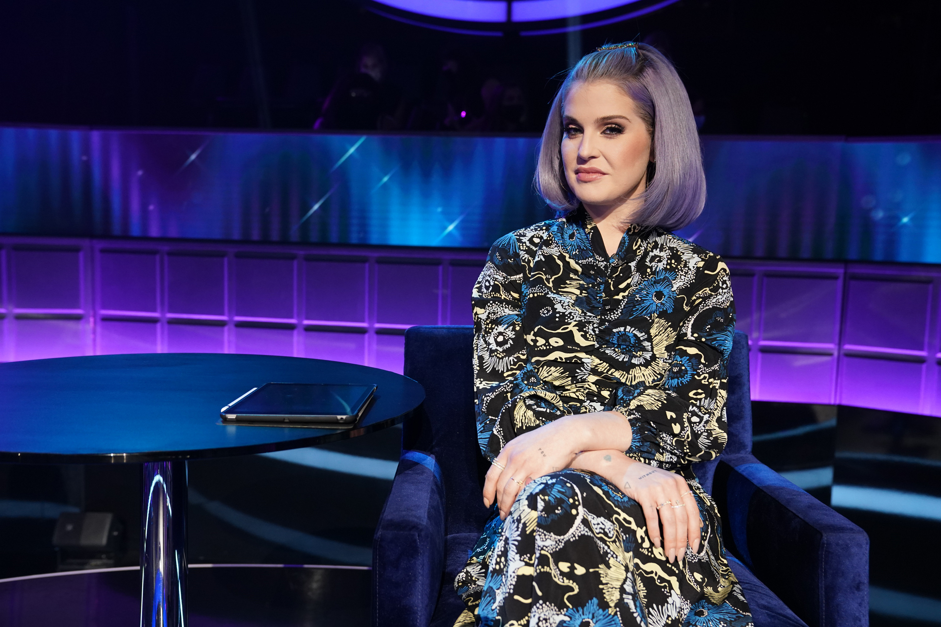 Kelly Osbourne seated on a TV show set, wearing a paisley-patterned outfit, styled hair