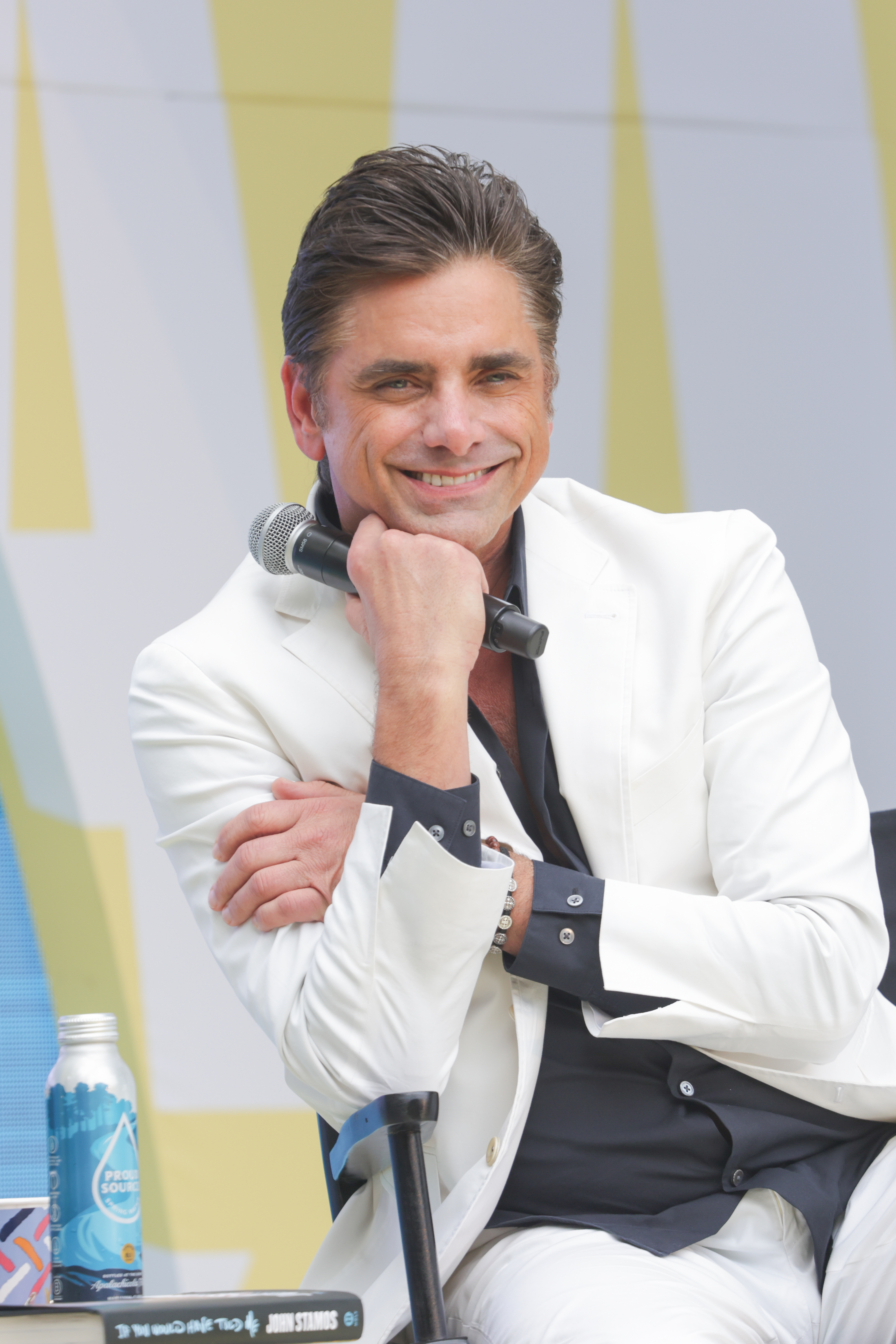 John Stamos wearing a white suit and vest, smiling while holding a microphone at an outdoor event