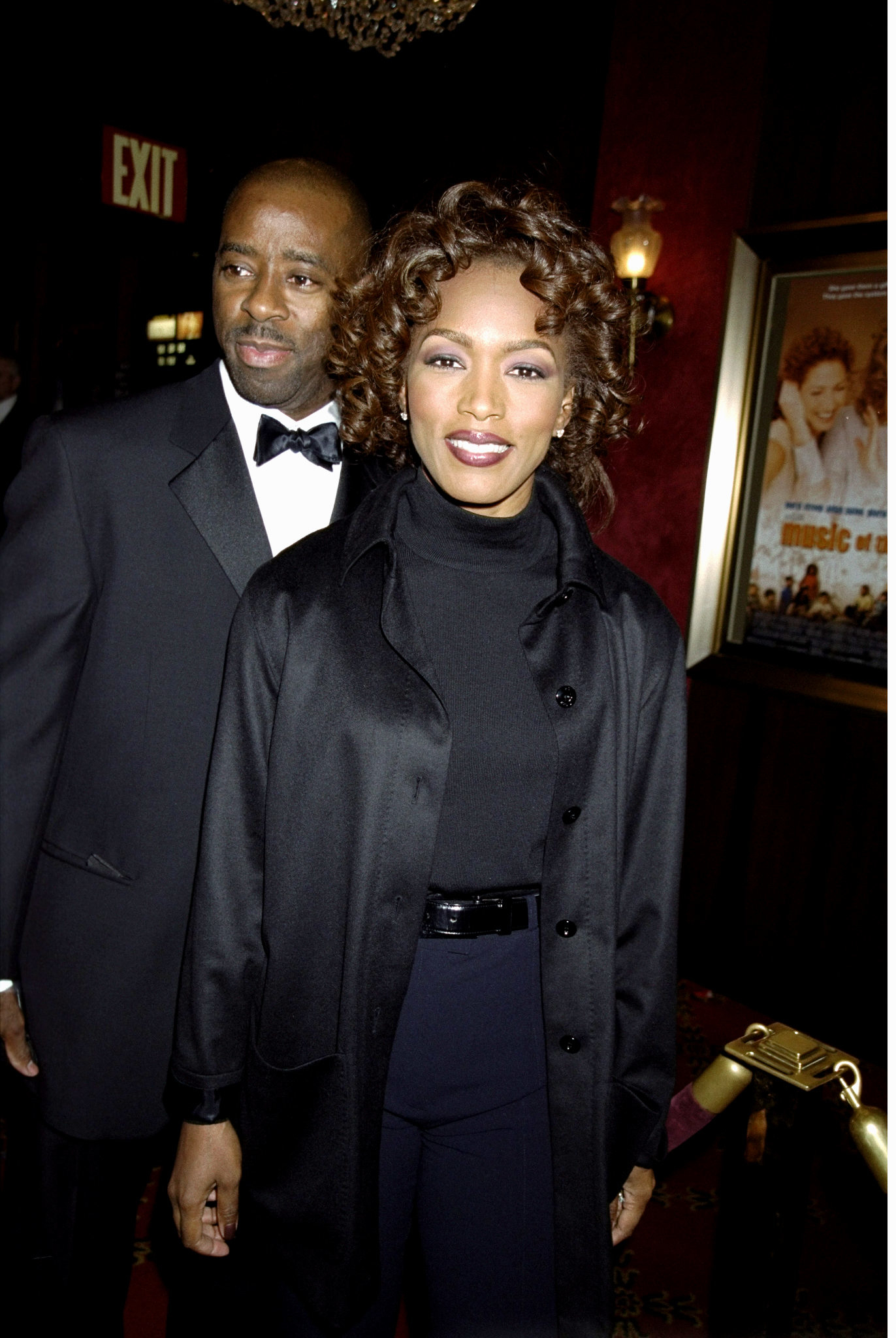 Two people posing together, one in a black suit and bowtie, and the other in a black coat and scarf