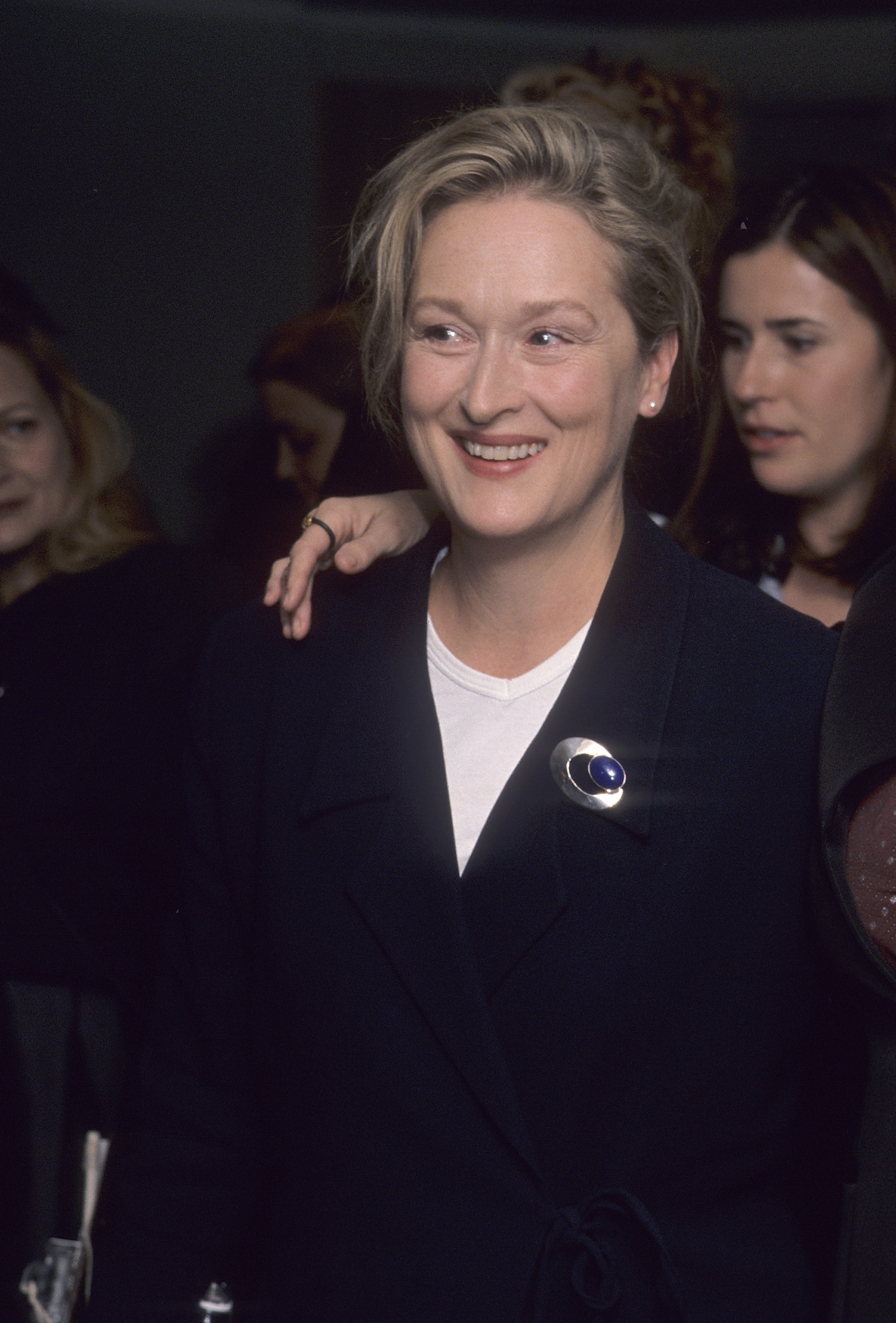 Meryl Streep in a black blazer with a round pin, smiling at an event