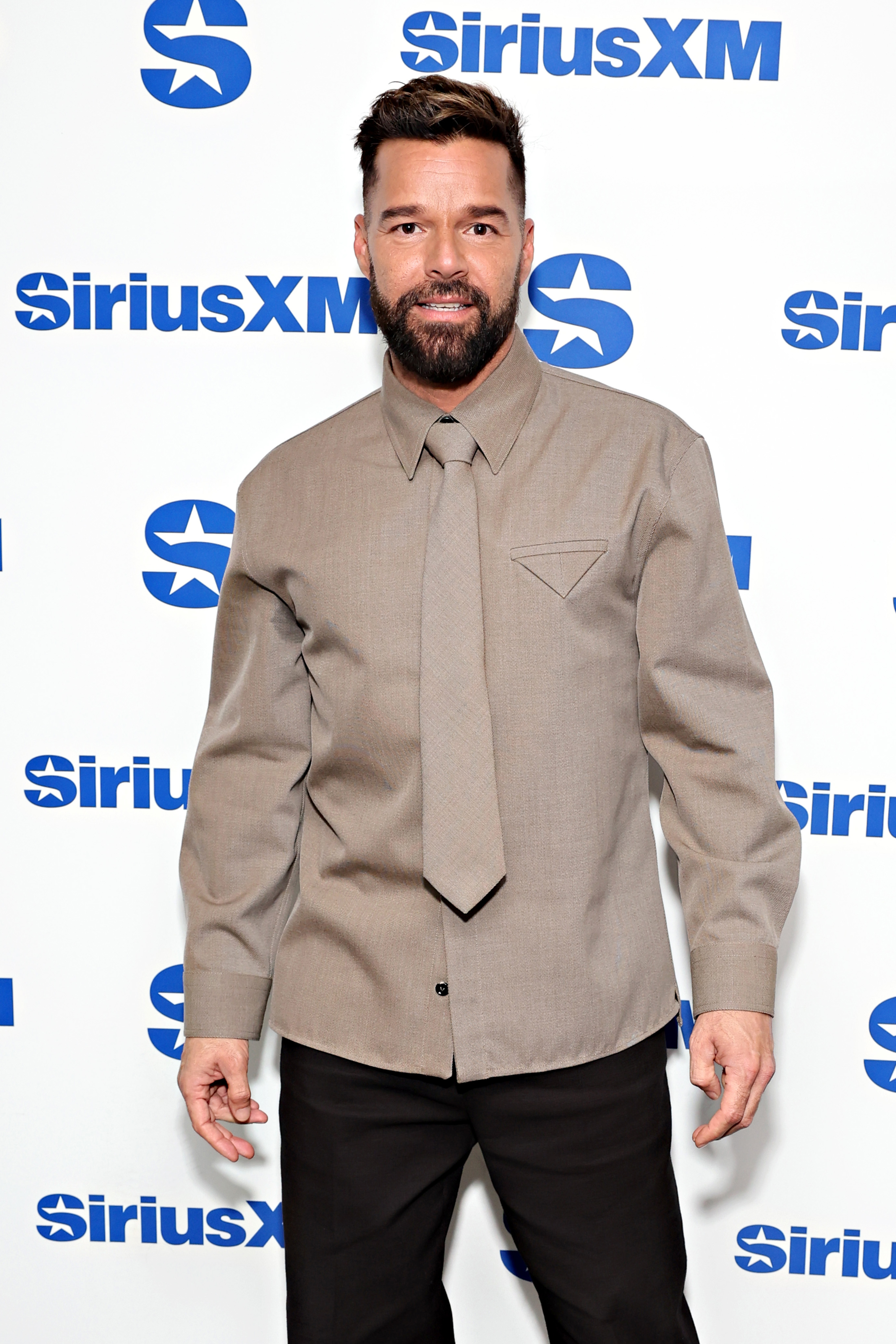 Ricky Martin poses in front of SiriusXM backdrop, wearing a stylish beige shirt and matching trousers