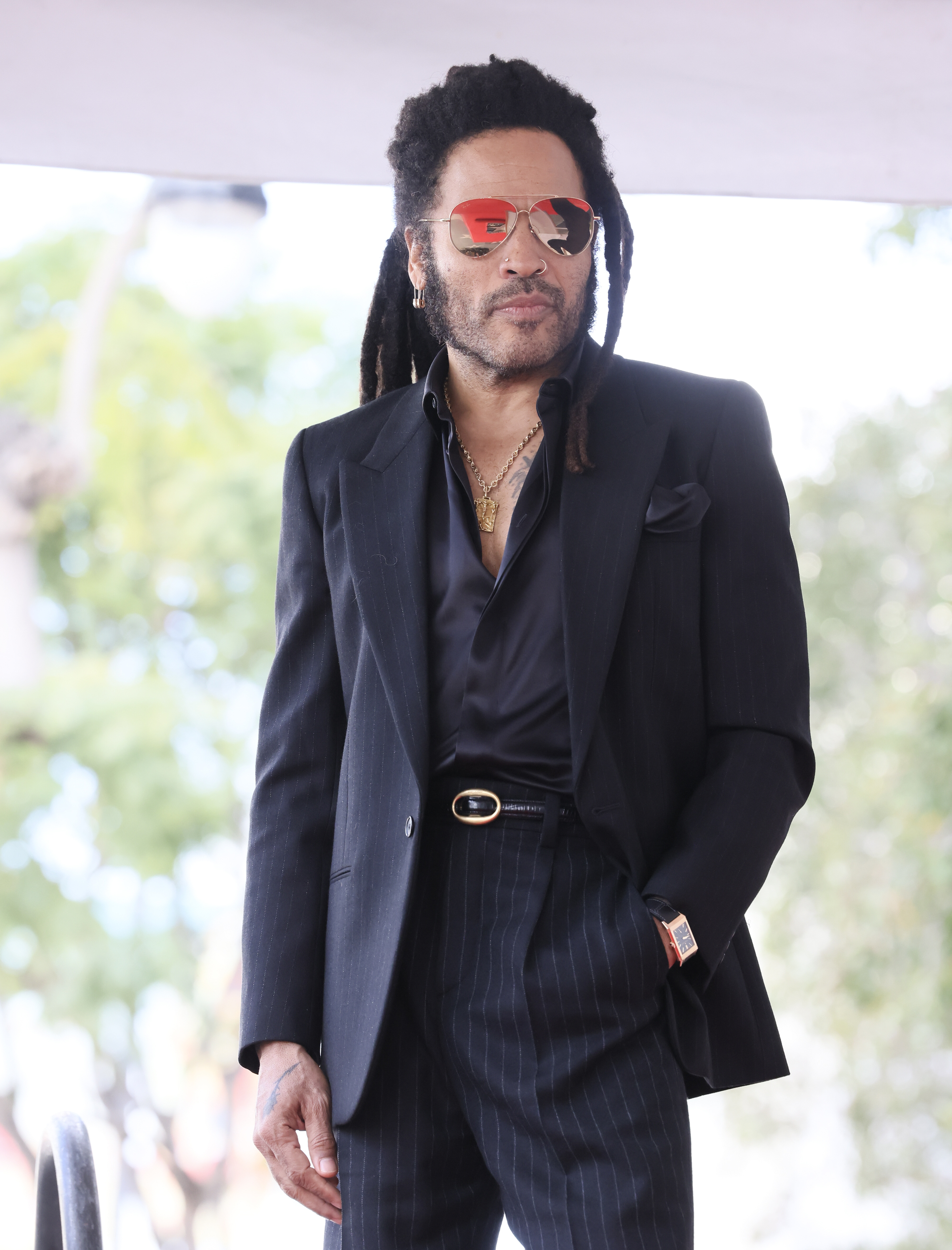 Man in a tailored suit with dreadlocks at an outdoor event, wearing sunglasses