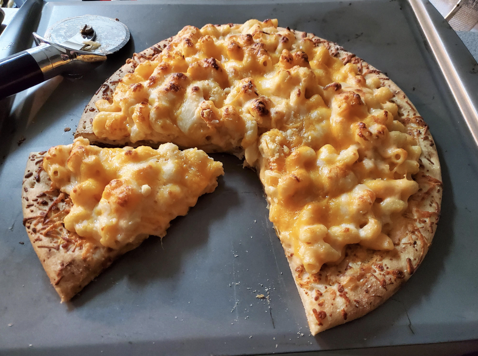 Pizza with macaroni and cheese topping, one slice removed, on a baking tray next to a pizza cutter