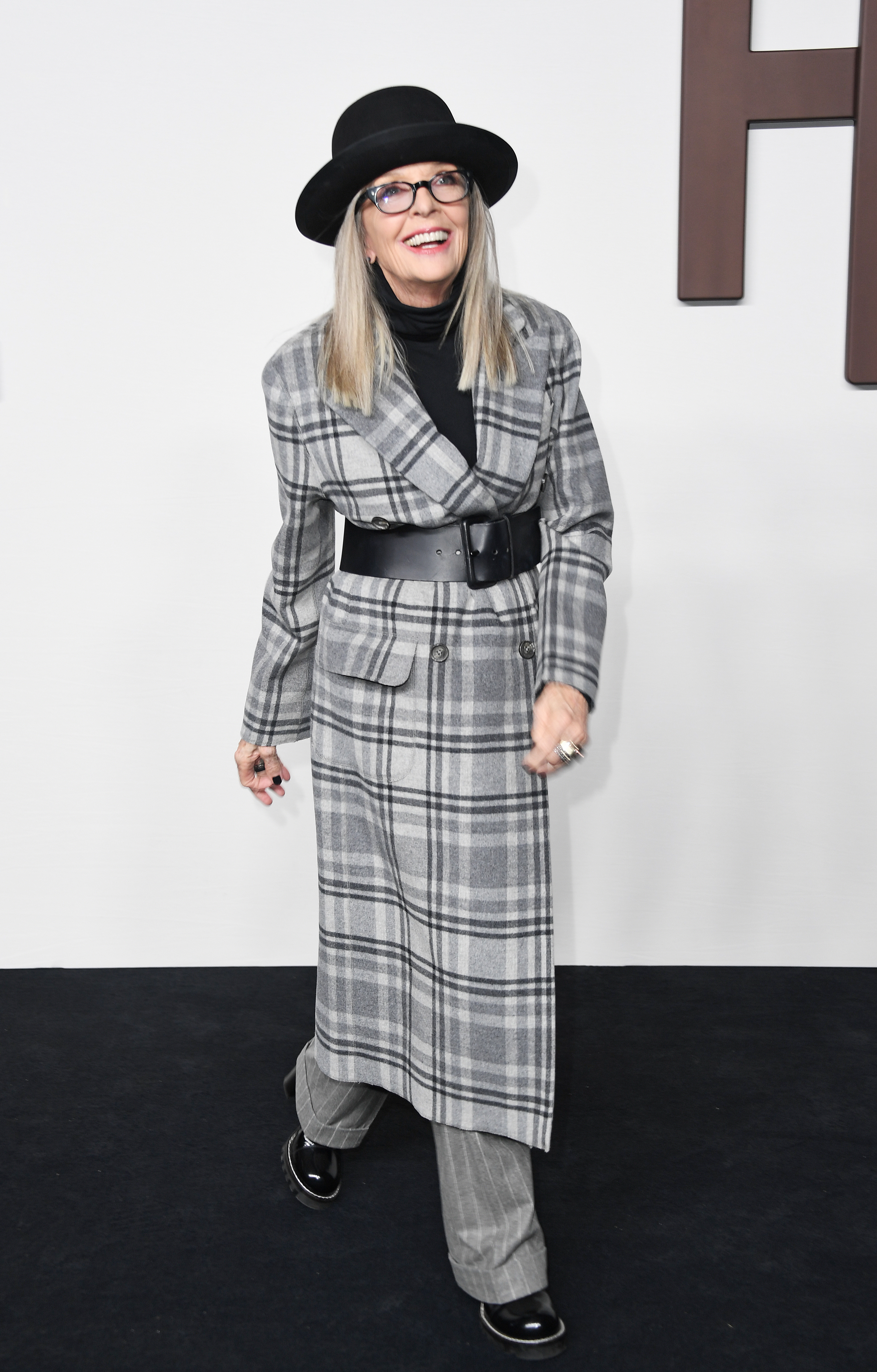 Diane Keaton in a plaid coat, turtleneck, and hat, poses with a smile at an event