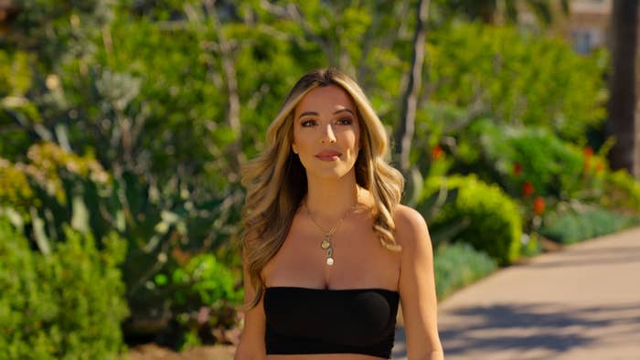 Alex in a strapless top with pendant necklace, standing on a garden path