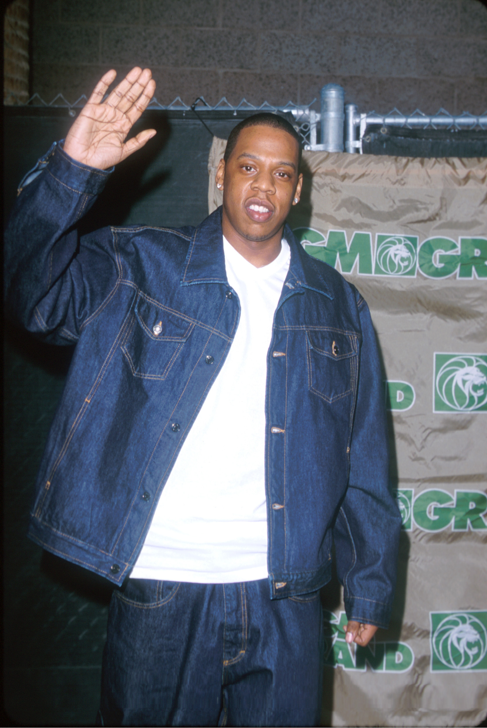 Jay-Z in a denim jacket, waving, with a backdrop featuring multiple logos