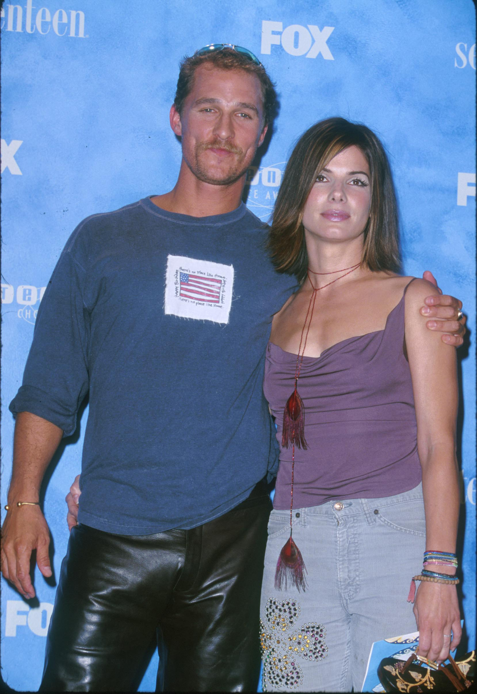 Two celebrities pose together; the man wears a casual shirt and leather pants, while the woman has on a tank top with a fringed detail