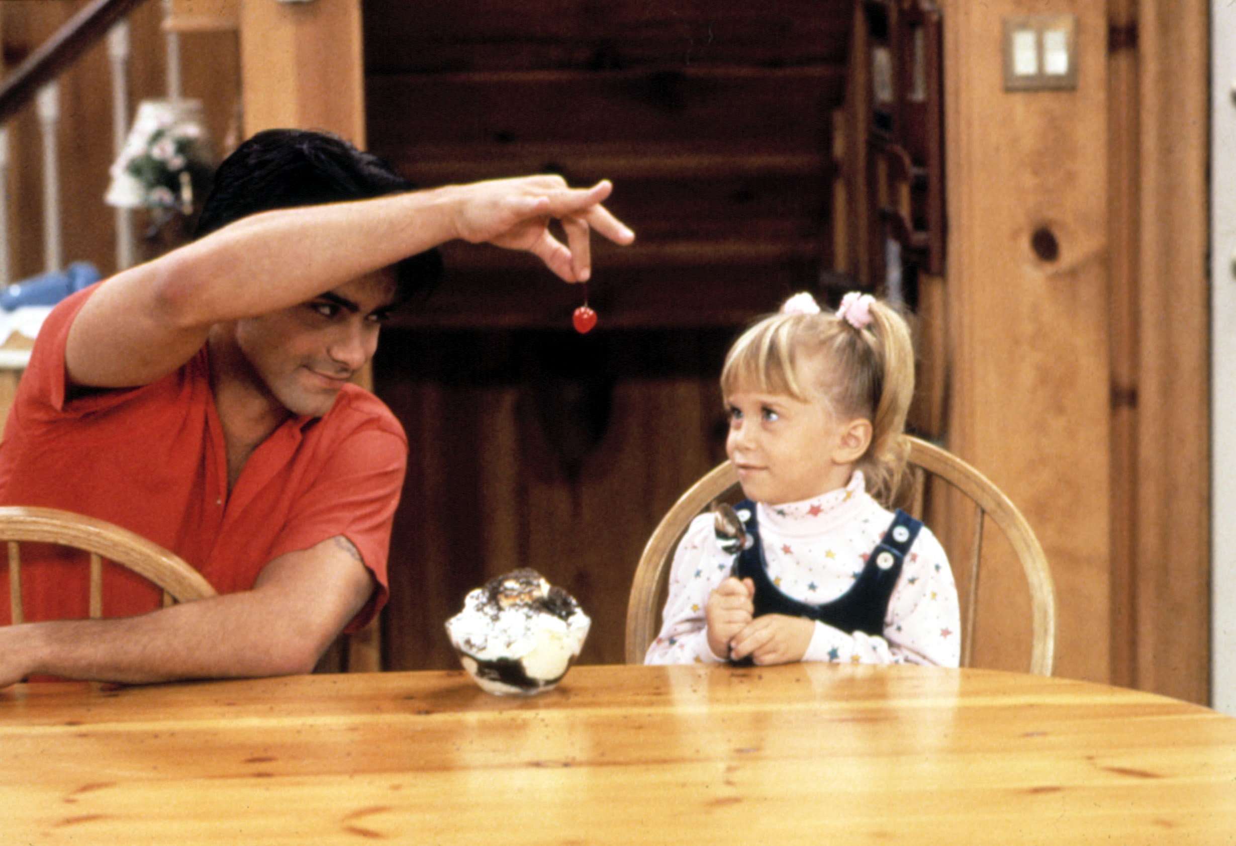 Uncle Jesse and Michelle from Full House sit at a table, with Jesse playfully dangling a cherry over Michelle&#x27;s head