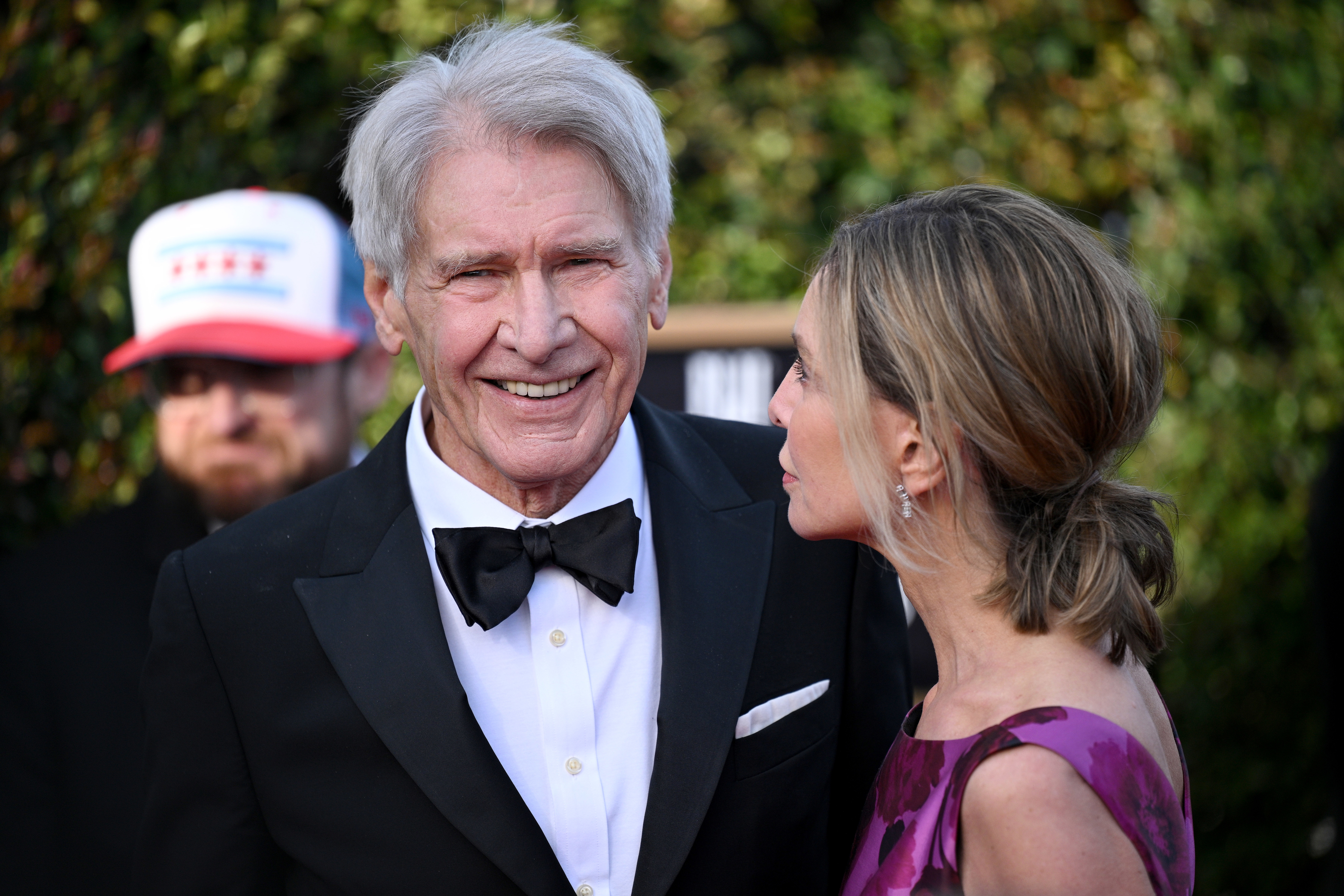 Harrison Ford in a black tuxedo with a bow tie shares a look with Calista Flockhart in a patterned gown